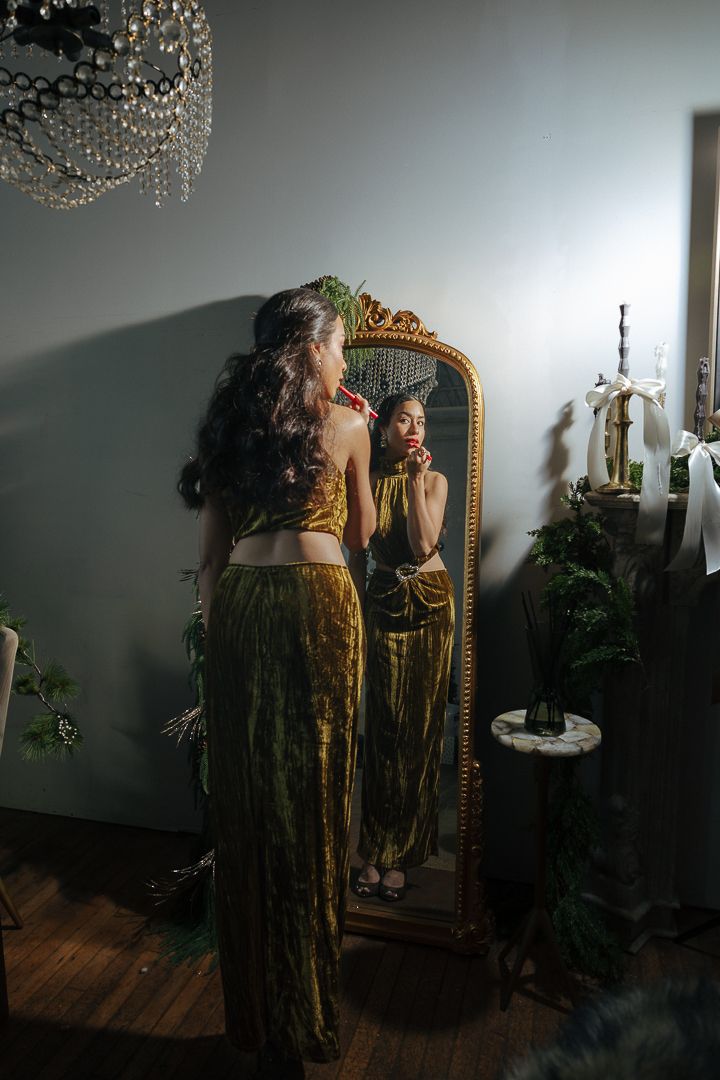 Woman in gold velvet two-piece outfit posing before ornate mirror in moody decorated room.