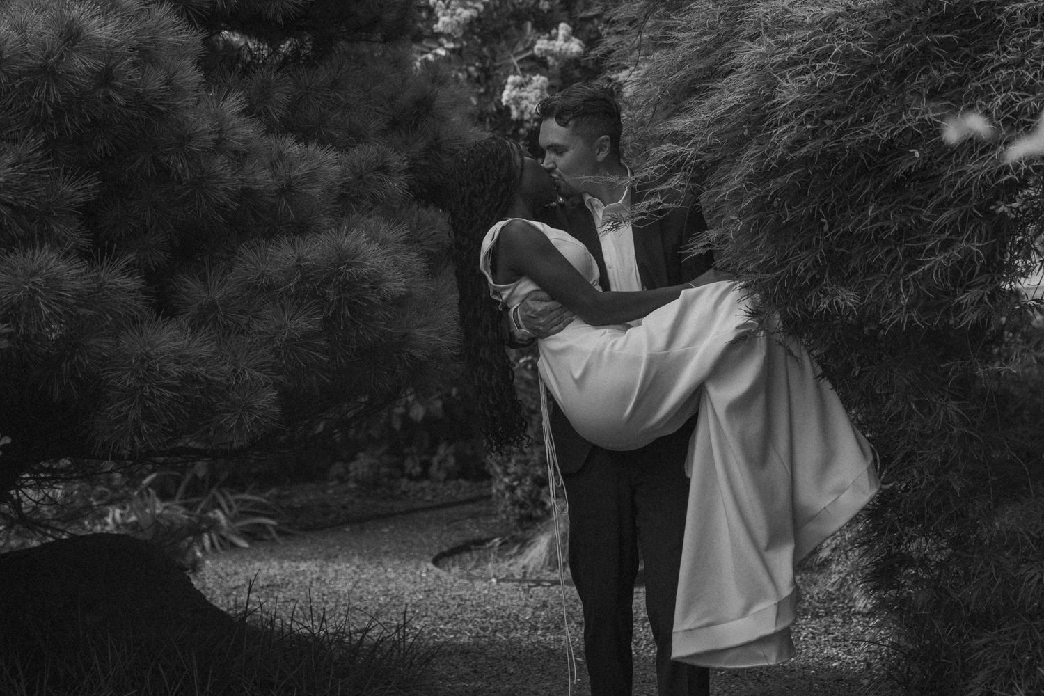 Black and white romantic photo of couple embracing in garden, groom lifting bride in flowing white dress.