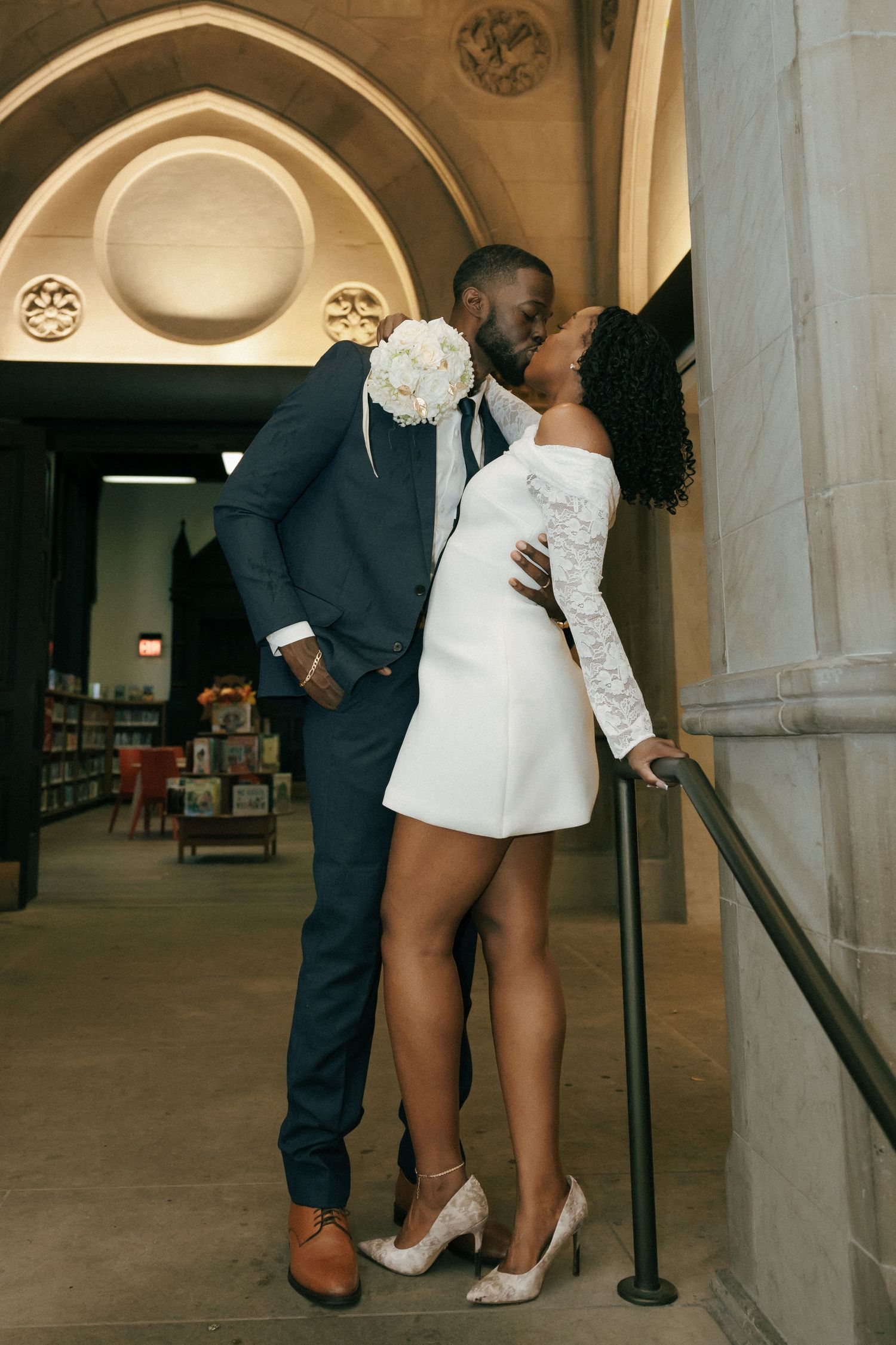 Bride and groom kissing in elegant arched hallway, bride in short white lace dress.