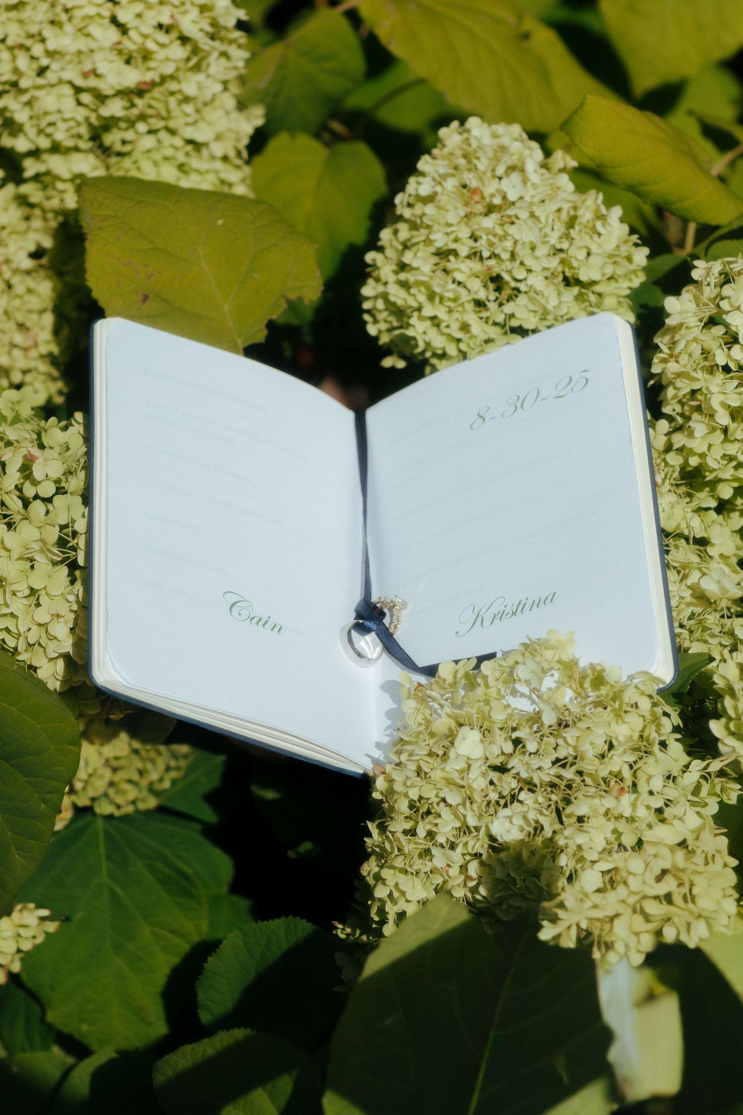 Open wedding journal with names Cain and Kristina dated 8.30.25 resting on white hydrangea blooms.