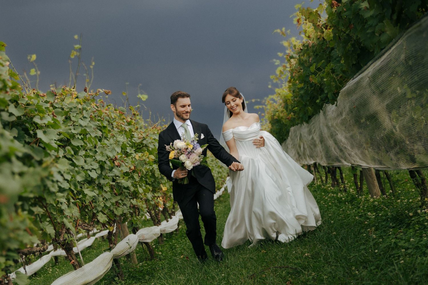 Smiling bride and groom walk hand in hand through a lush vineyard under a dramatic stormy sky.