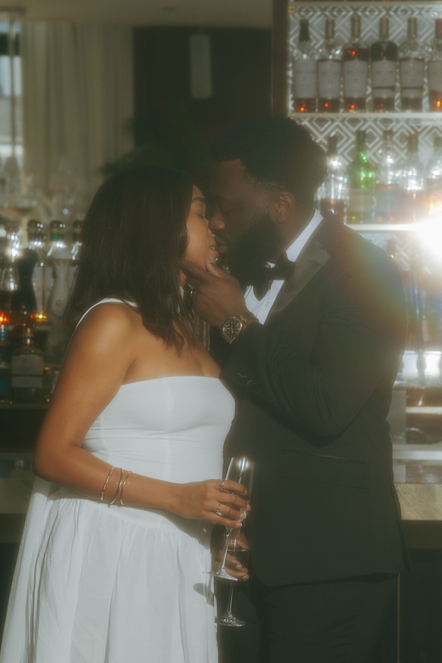 A couple shares a romantic kiss at a bar, woman in white strapless dress holding champagne, man in black tuxedo.
