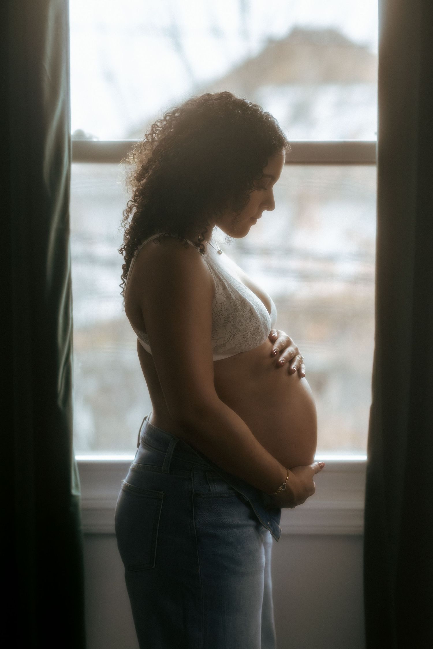 Pregnant woman in white bralette and jeans cradles her baby bump while standing by a softly lit window.