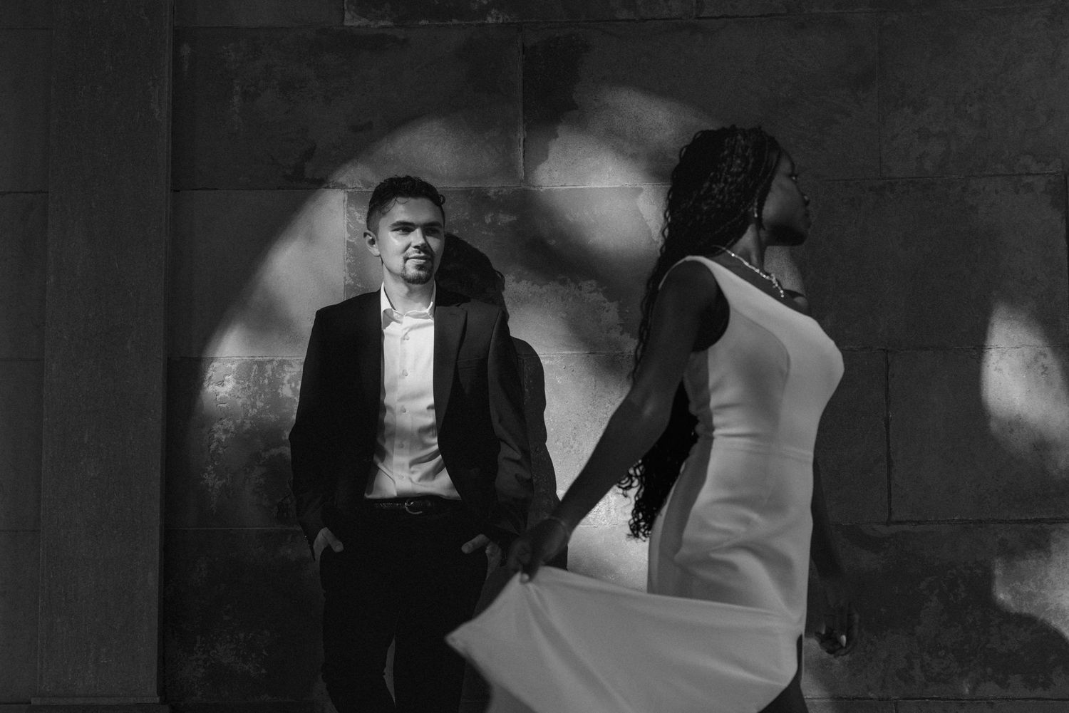 Black and white photo of a couple in formal wear, man in suit and woman in white dress, standing by a stone wall.