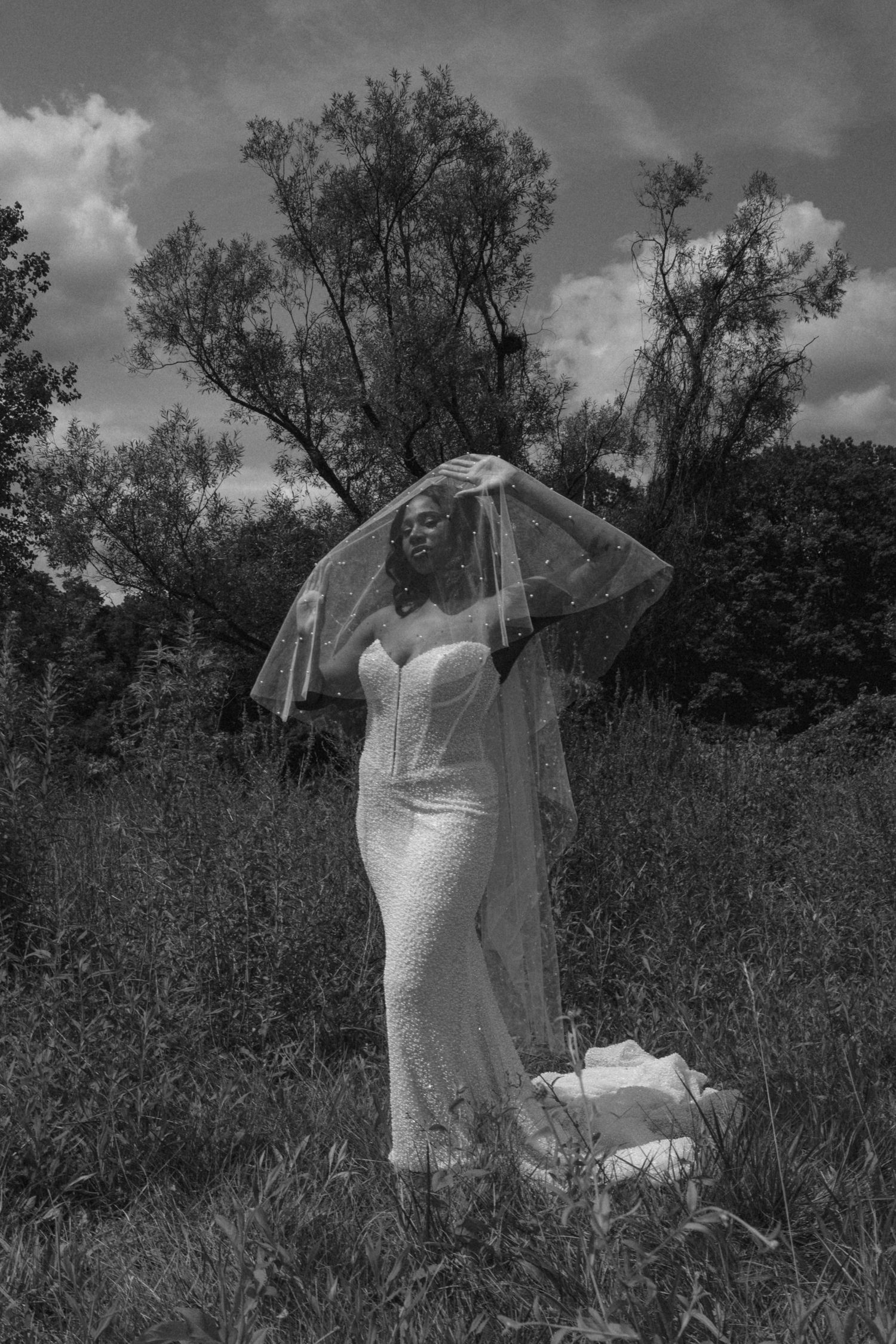 Black and white photo of bride in fitted lace gown lifting veil in wild meadow.