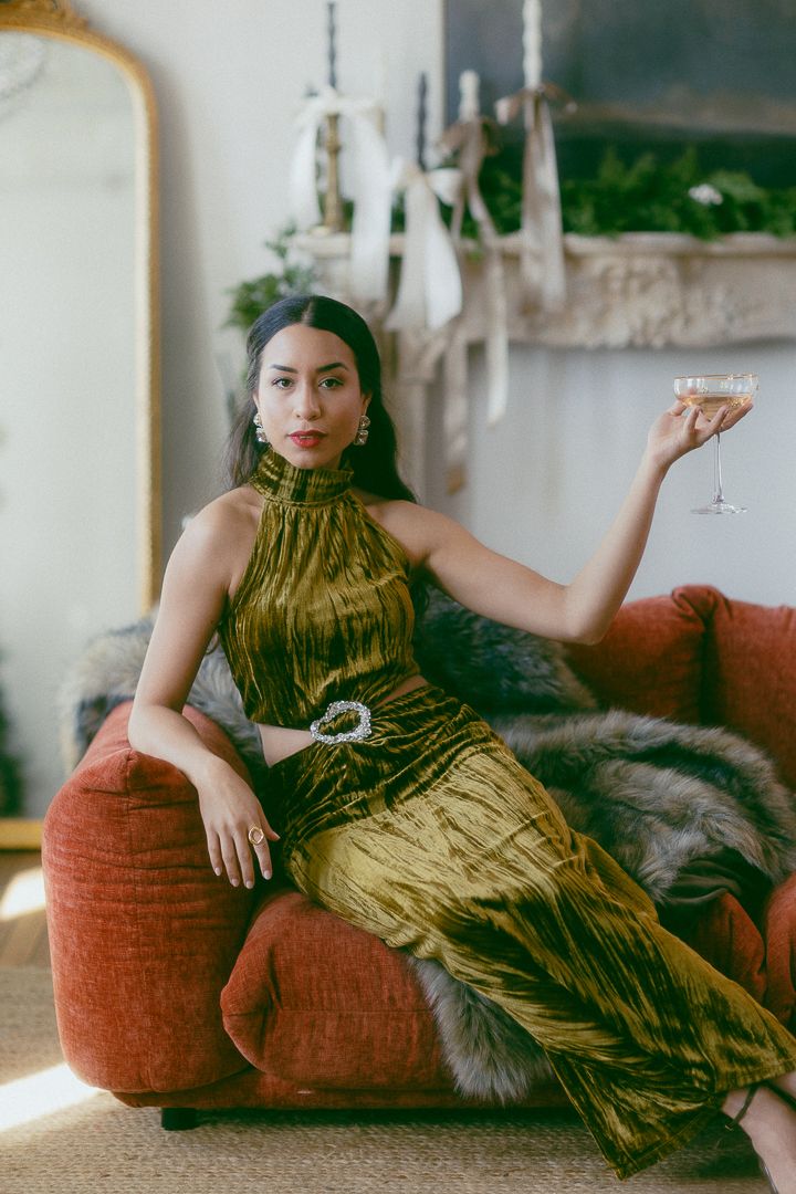 Woman in gold velvet halter dress sitting on red sofa holding champagne glass in elegant holiday interior.