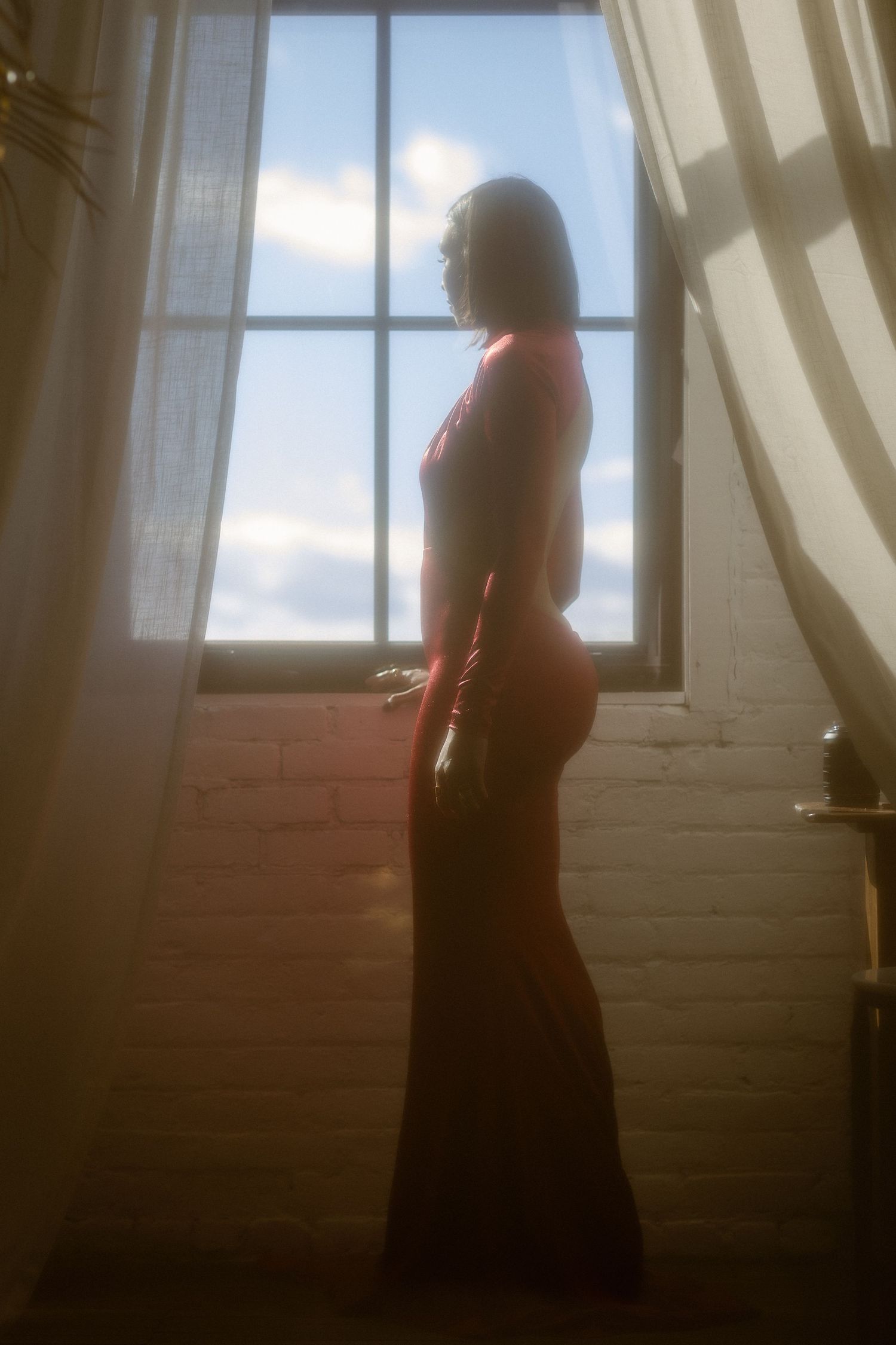 Silhouette of a woman in a red dress standing by a sunlit window with sheer curtains.