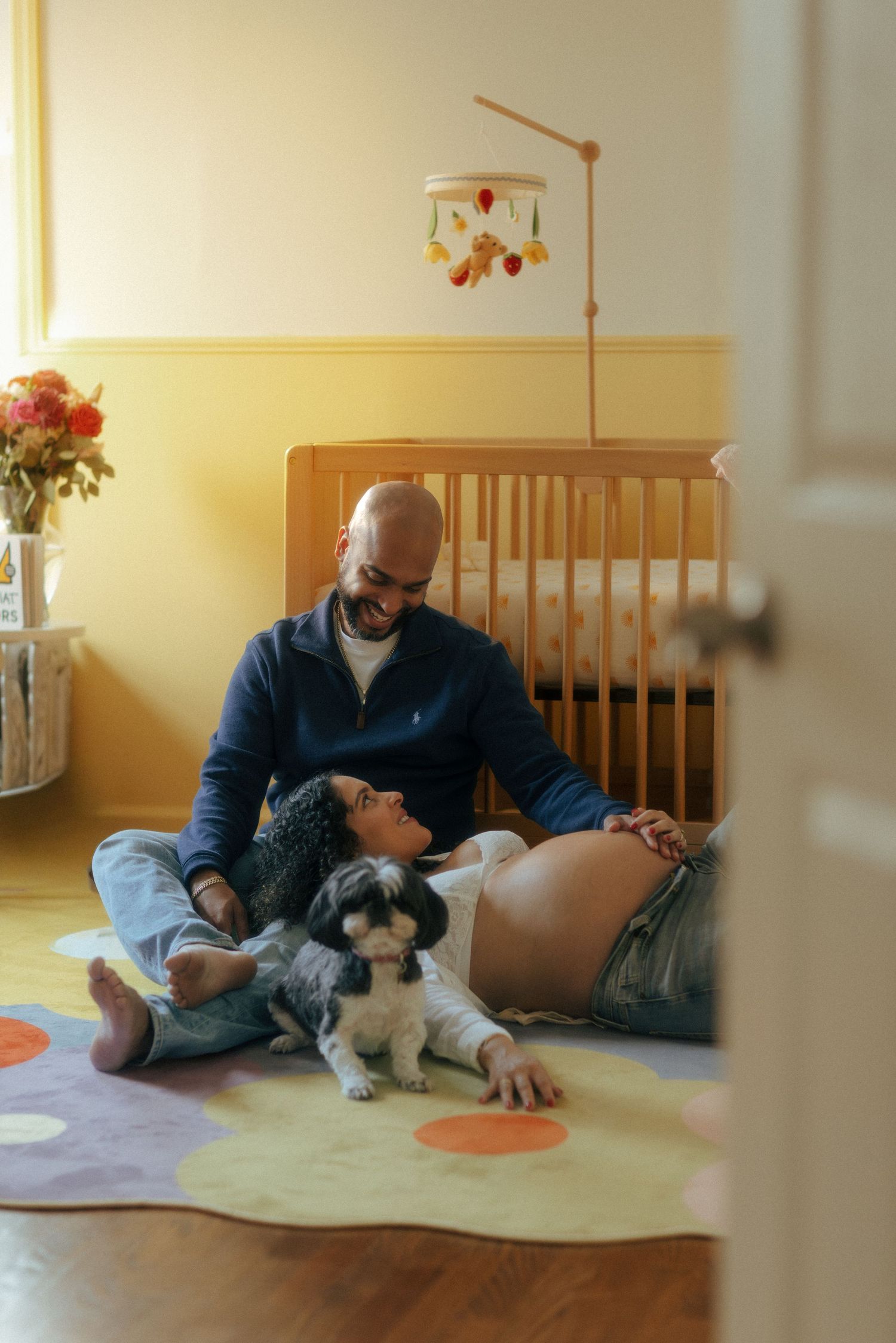 Happy couple with dog relaxing on nursery floor beside wooden crib in warm yellow room.