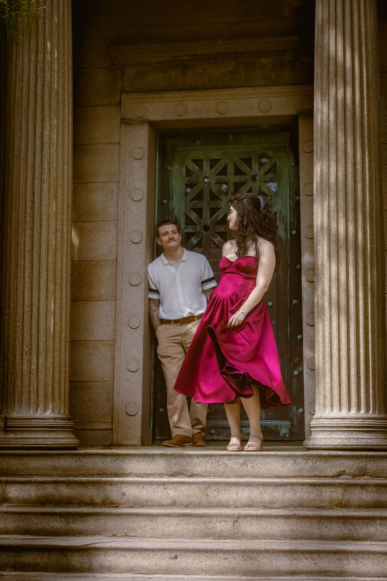 Couple posing on stone steps between columns, woman in flowing red dress, man in white shirt, elegant architectural backdrop.