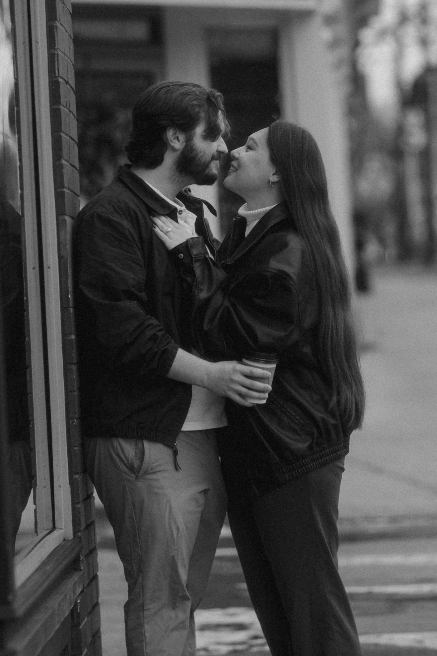 Black and white photo of a romantic couple about to kiss on a city sidewalk, both wearing dark jackets.