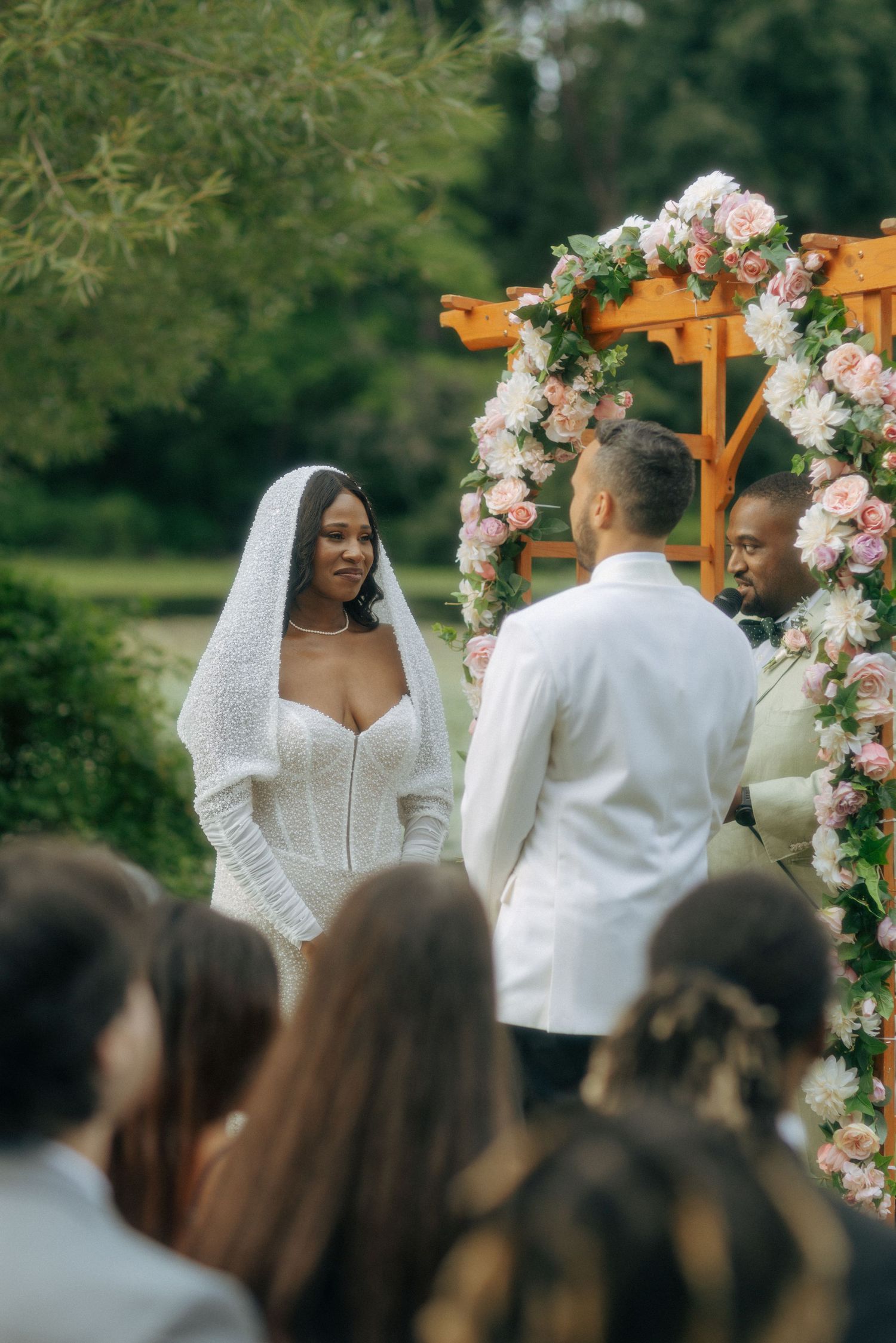 A bride in a white knit gown and veil exchanges vows under a floral arch at an outdoor ceremony.