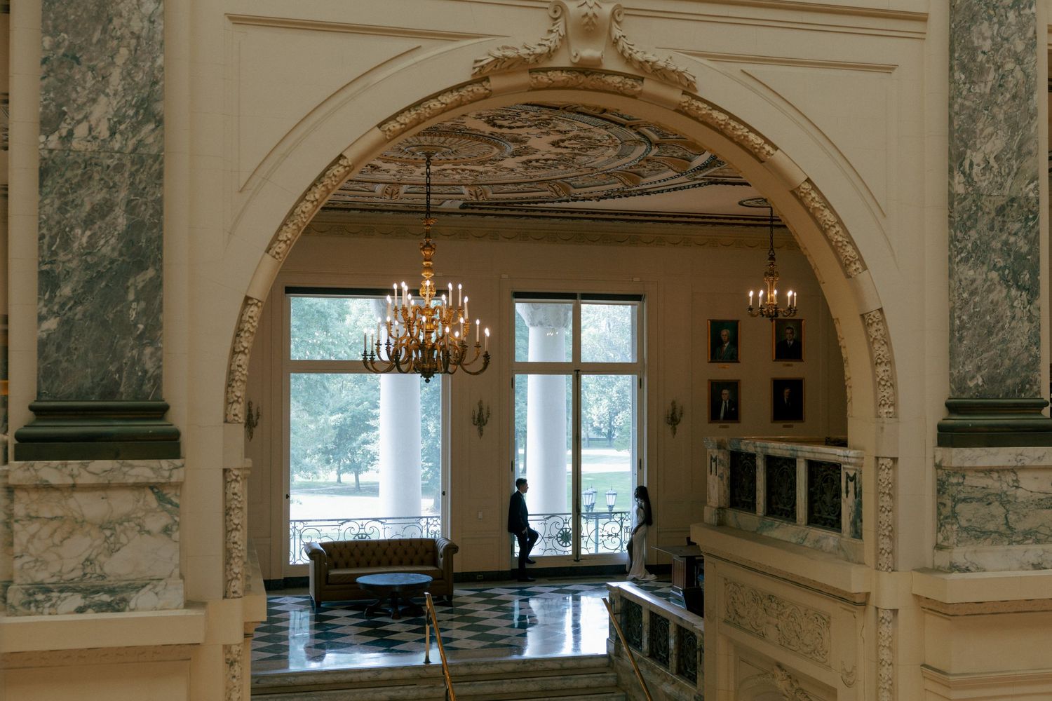 Elegant grand hall interior featuring ornate white archway, chandelier, marble columns, and decorative painted ceiling.
