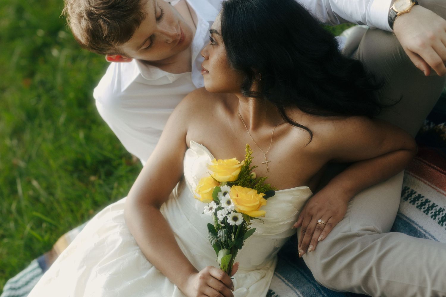 Bride and groom sitting on grass, bride holding yellow roses bouquet in a white strapless wedding dress.