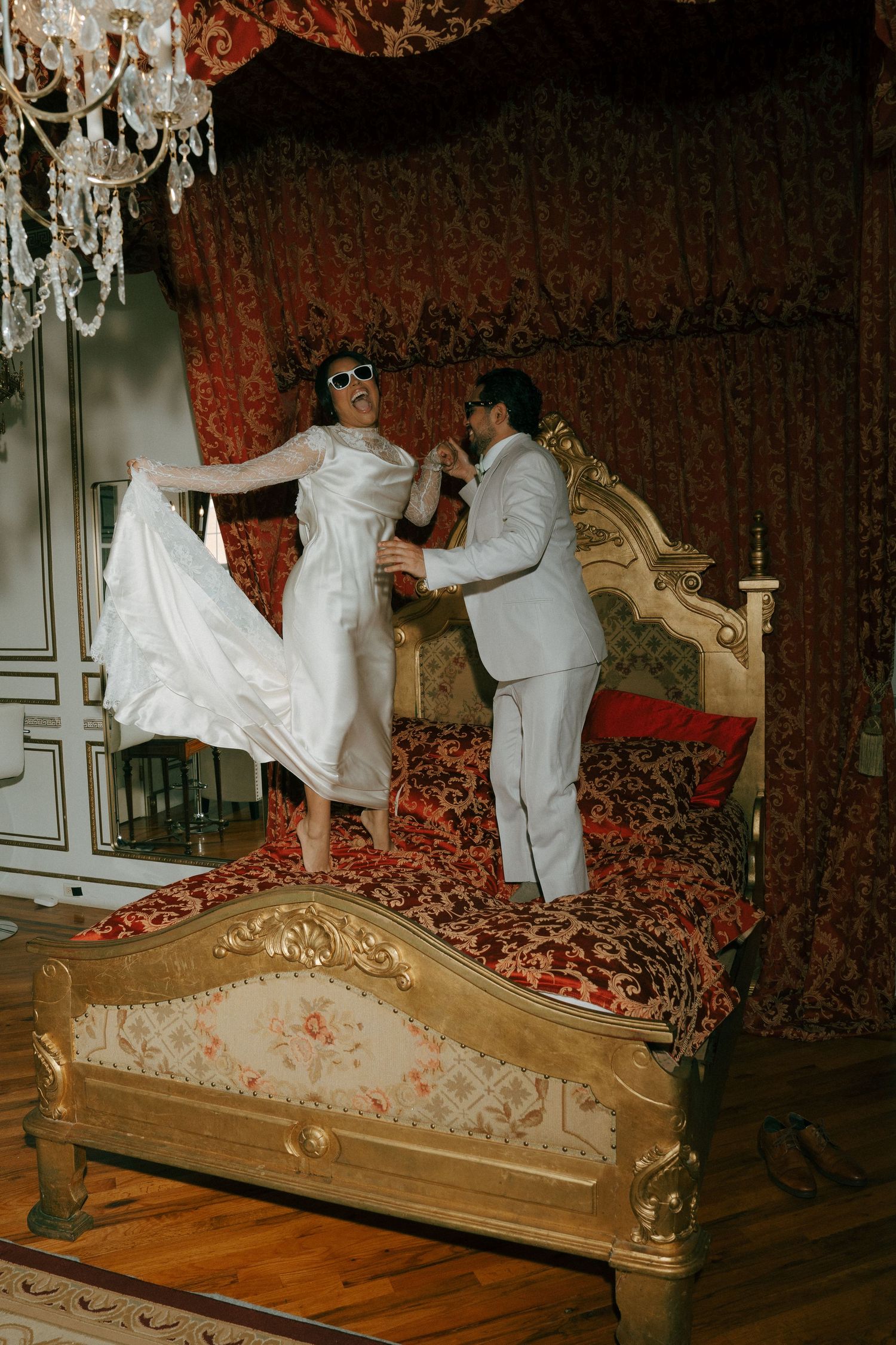 Bride and groom in sunglasses dancing on an ornate golden bed in a luxurious red-draped vintage bedroom.