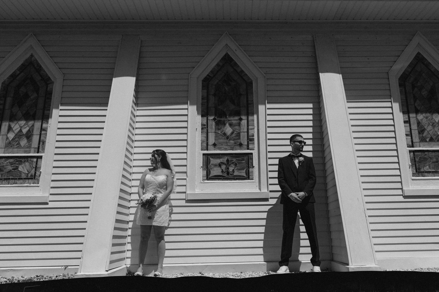 Black and white photo of a bride and groom standing apart outside a white church with Gothic stained glass windows.