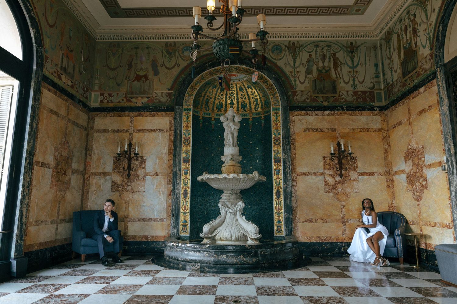Bride and groom posed apart in an ornate baroque room with a decorative fountain and painted walls.