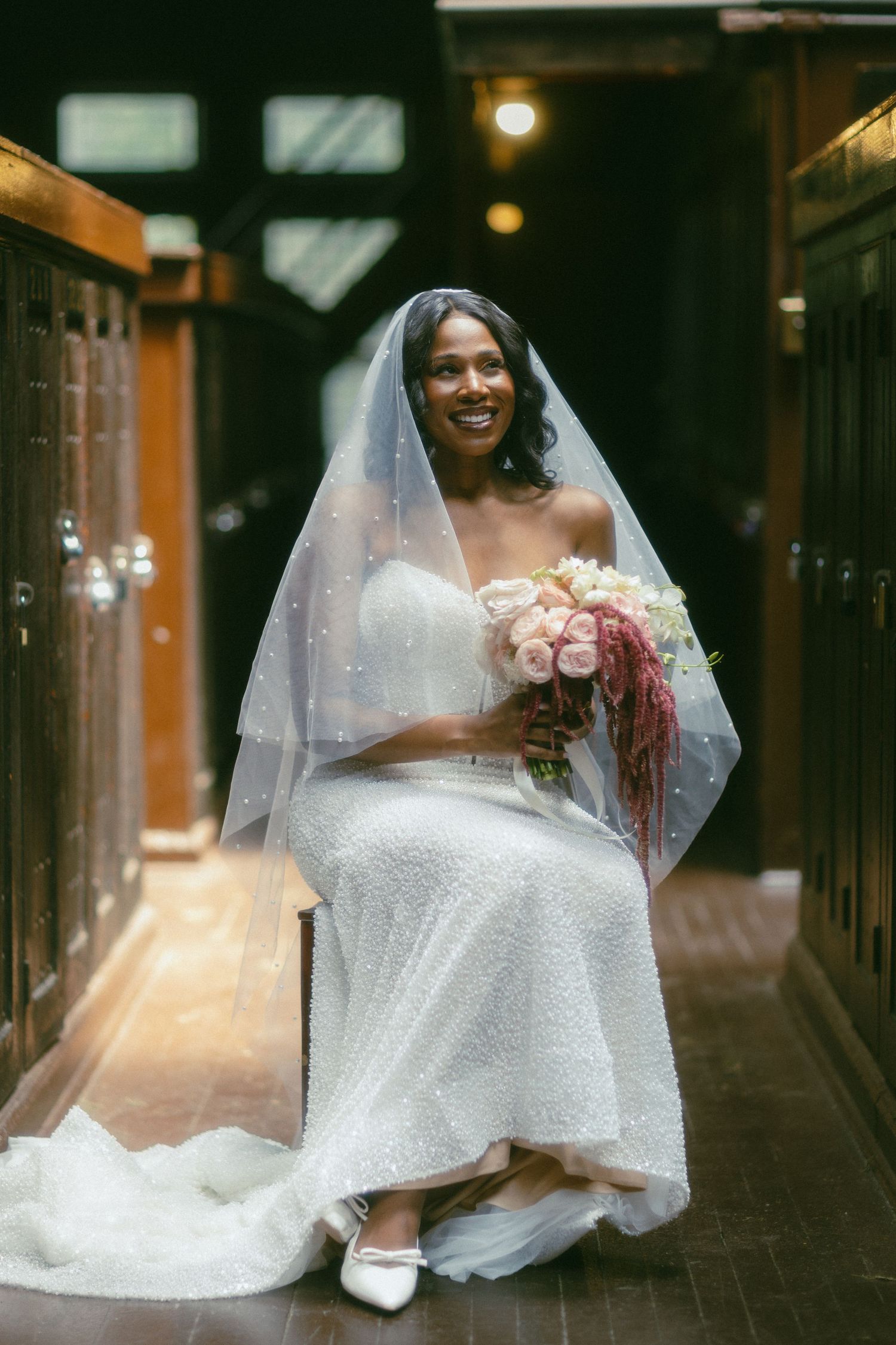 Smiling Black bride in white lace gown and veil sits holding pink bouquet in elegant indoor corridor.