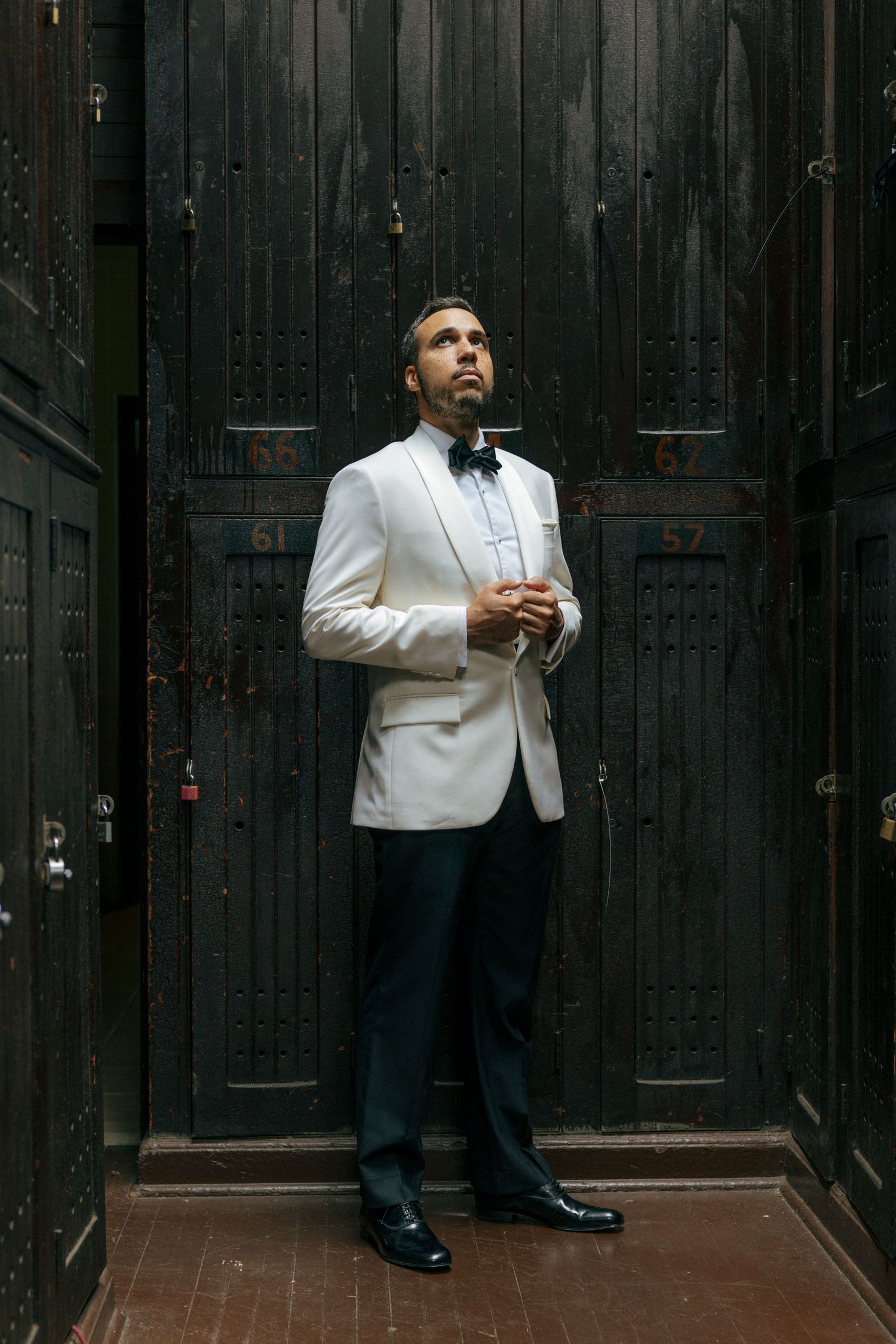 Groom in white tuxedo jacket and black bow tie standing before dark wooden lockers.