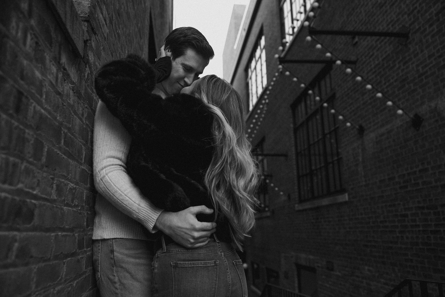 A couple shares a romantic embrace in a moody black and white alley with string lights overhead.