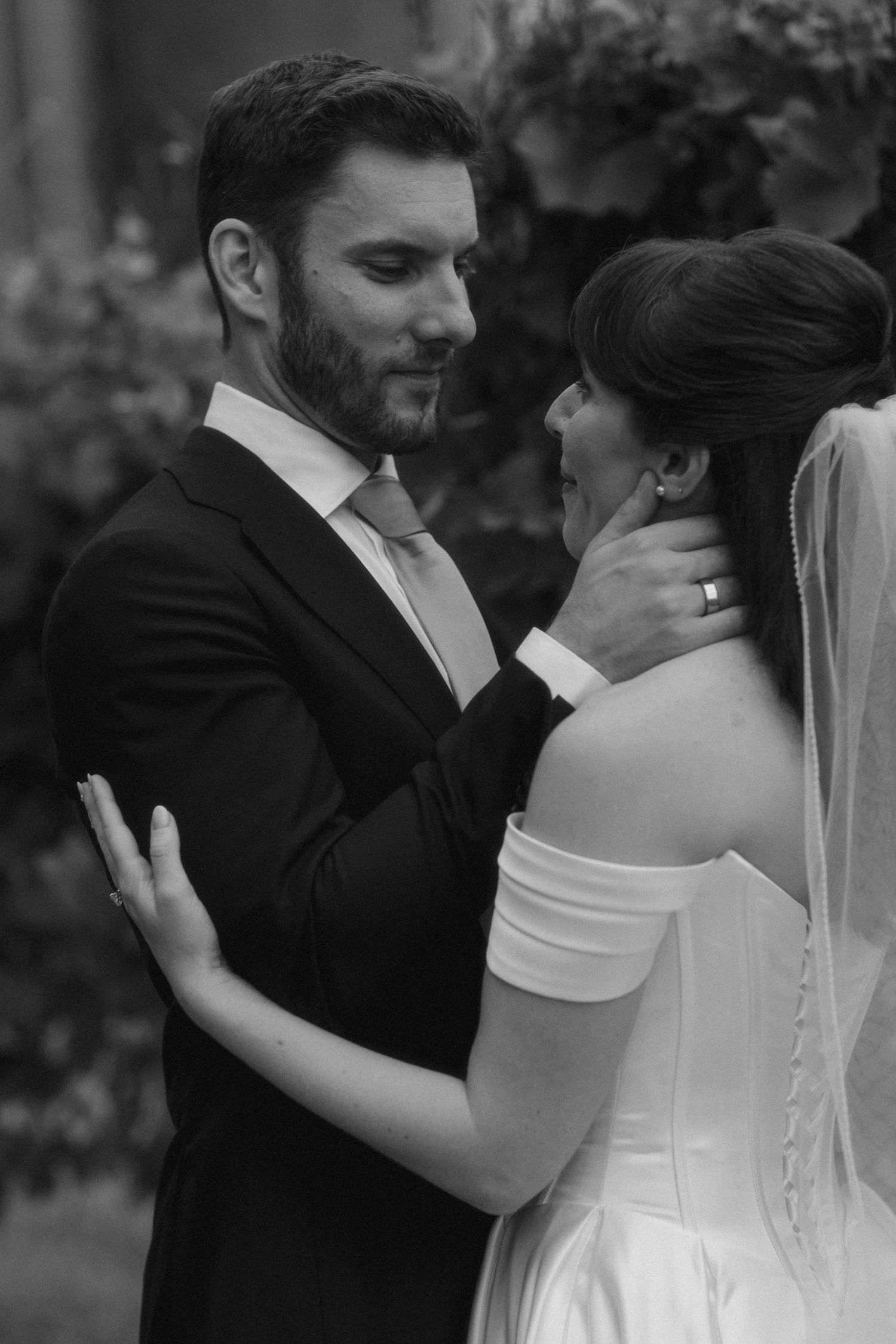 Black and white photo of a groom tenderly holding his bride's face as they share an intimate moment outdoors.