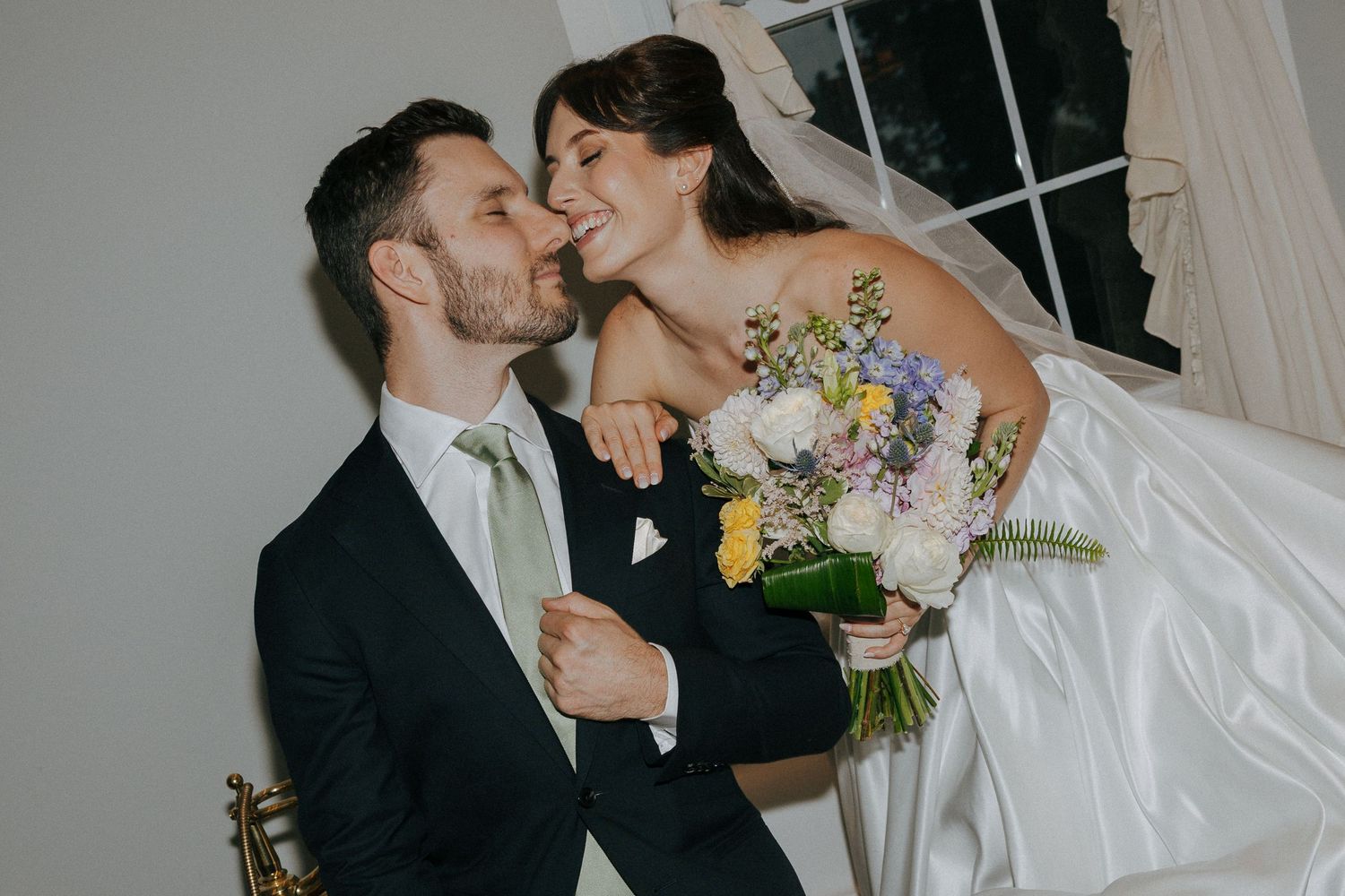 Joyful bride leans down to embrace groom, laughing together while holding a pastel wildflower wedding bouquet indoors.