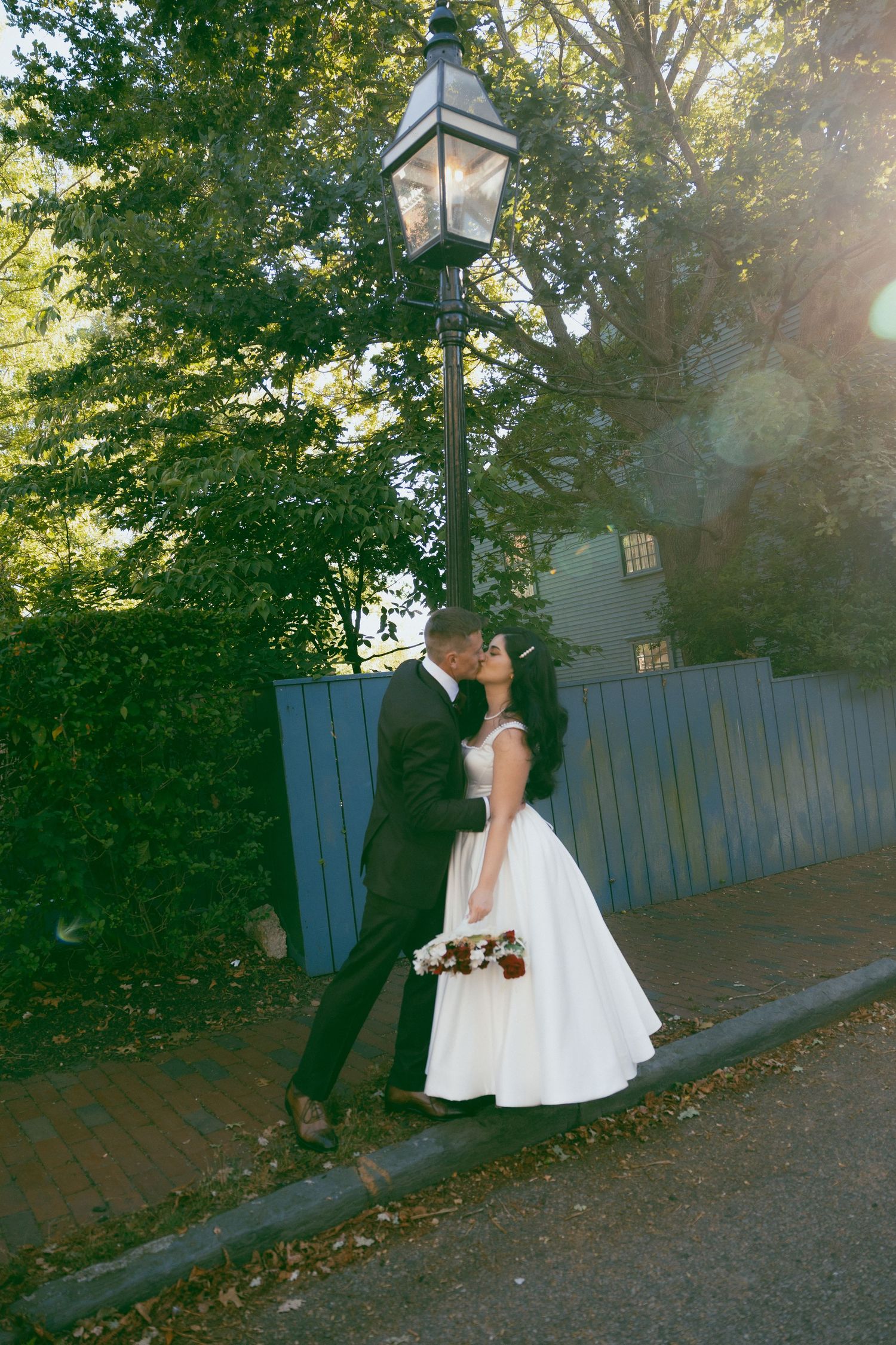 Bride and groom kissing under vintage street lamp surrounded by lush green trees in golden sunlight.
