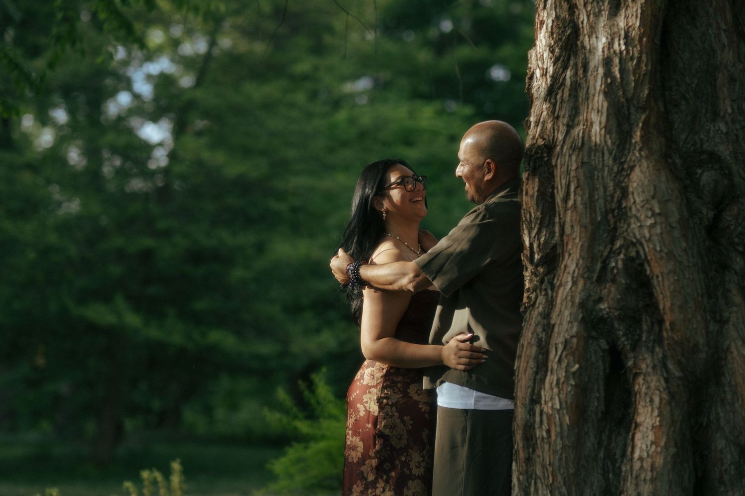A couple embraces romantically near a large tree in a lush green park setting.