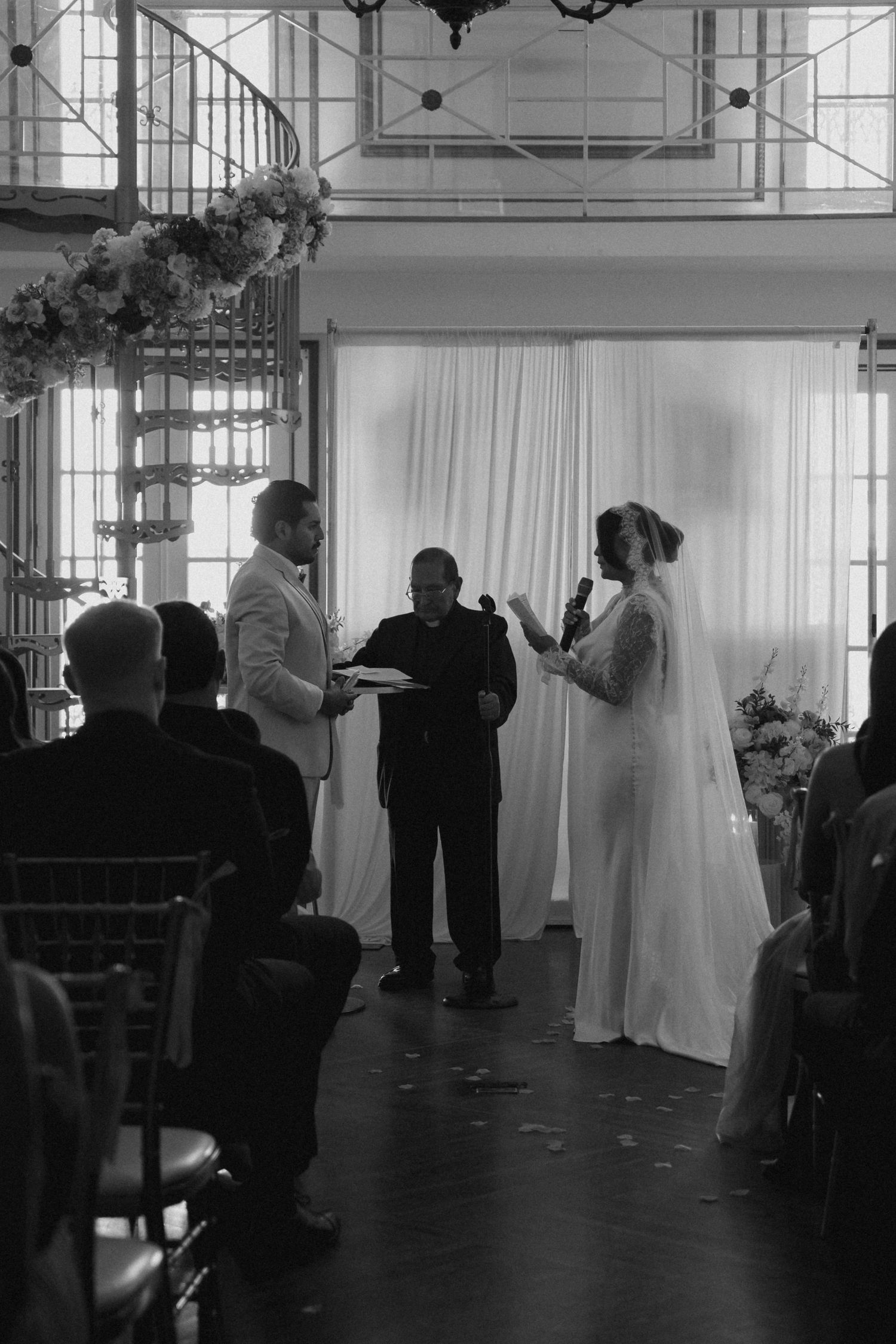Black and white photo of a bride reading vows at an indoor wedding ceremony with an officiant and groom.