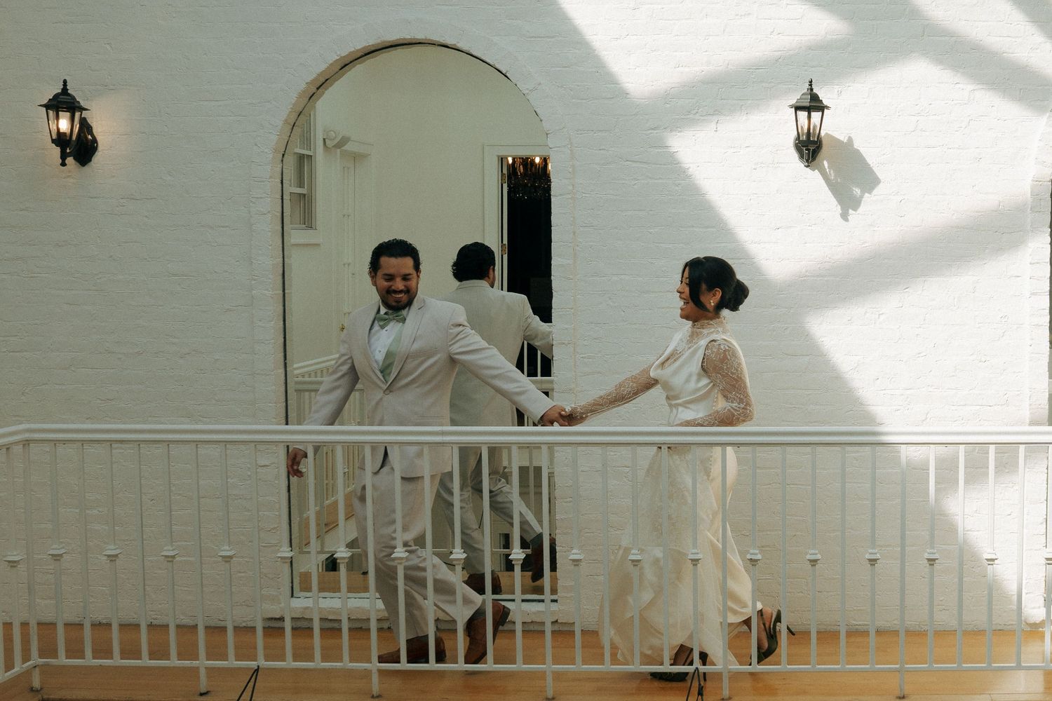 Bride and groom hold hands on a white balcony outside an arched doorway at an elegant wedding venue.