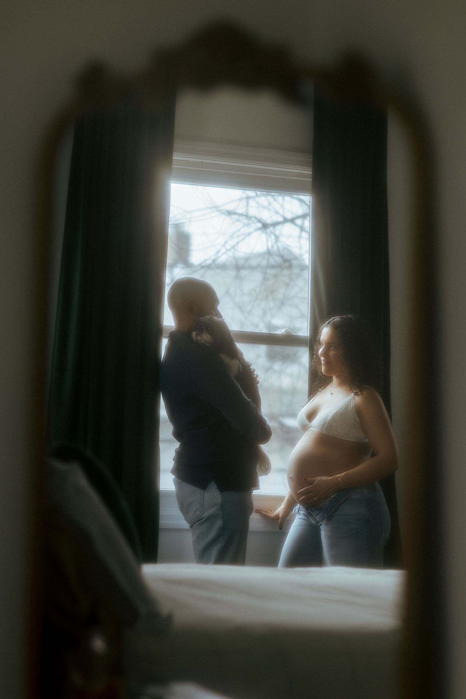 Couple sharing a tender moment by a bright window, reflected in an ornate mirror, with pregnant belly visible.