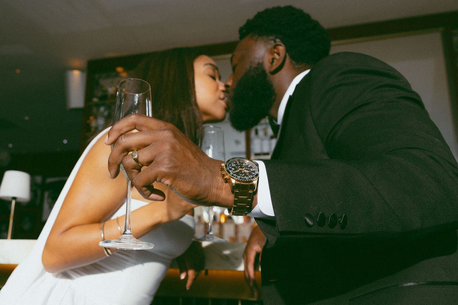 A couple in formal attire shares a romantic kiss while holding champagne glasses at a wedding reception.