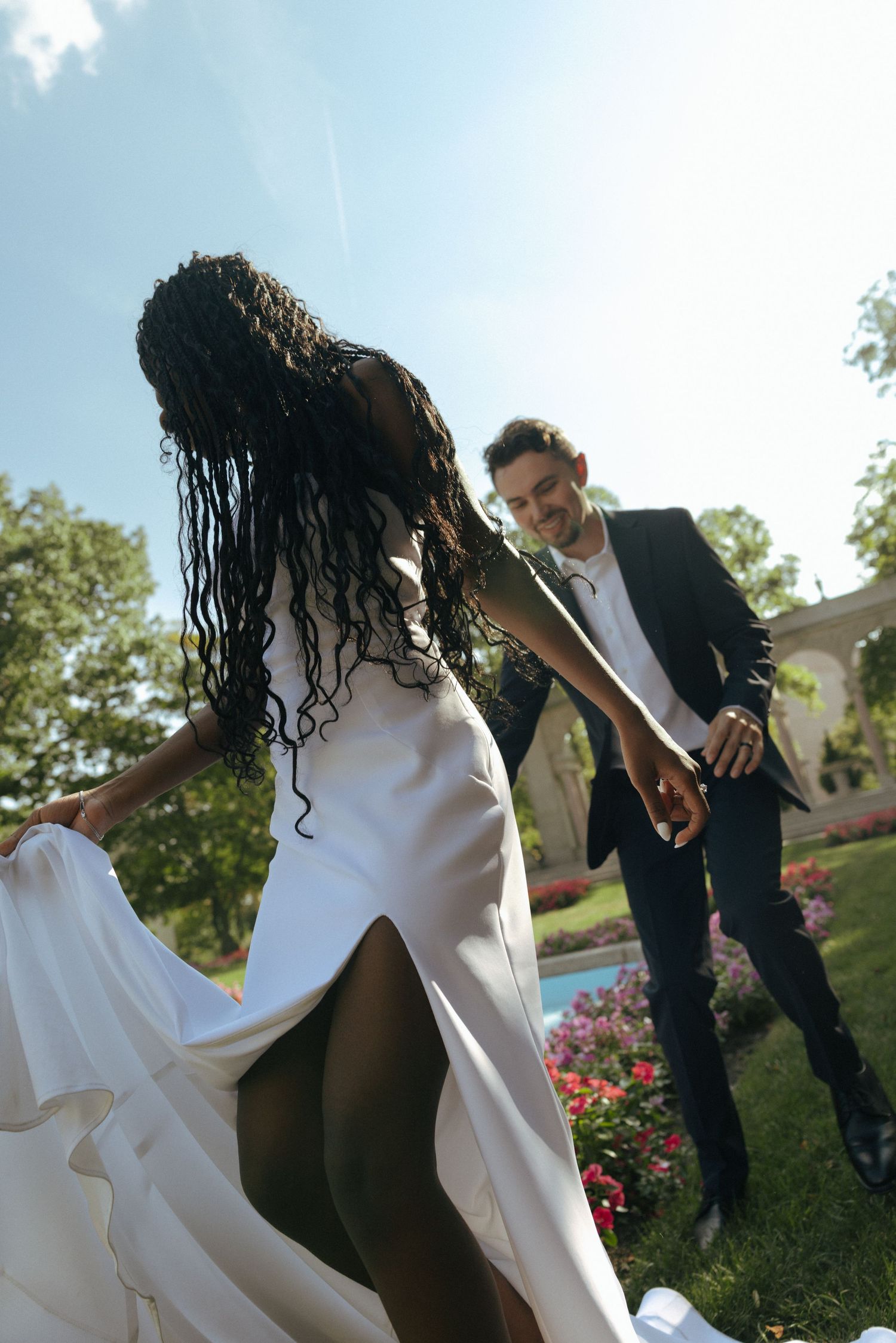 Interracial couple in wedding attire dancing outdoors, bride in white gown holding dress, groom in black suit smiling.
