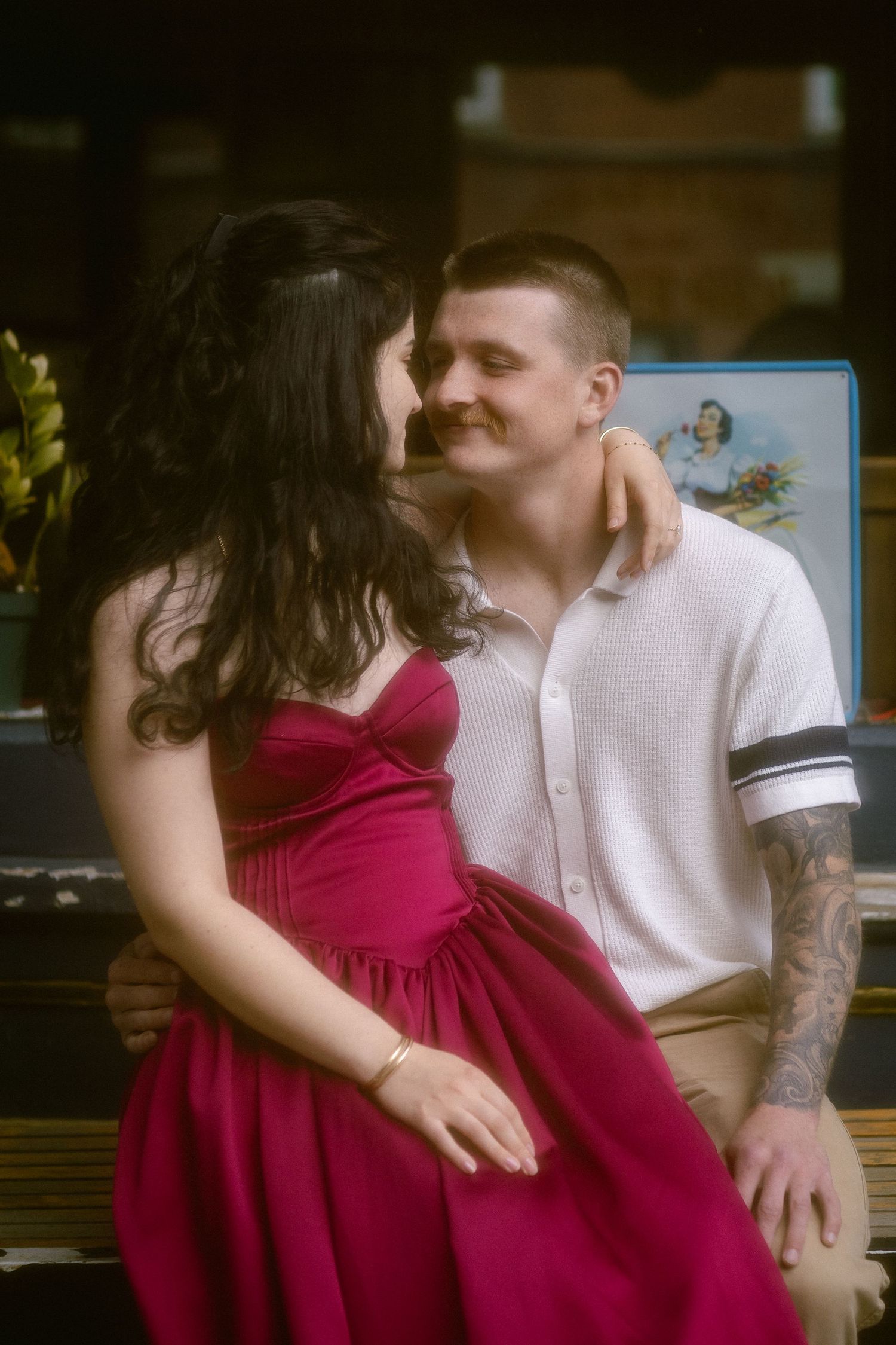 A couple shares a romantic moment on a bench, woman in red dress with dark hair, man in white polo with tattoos.