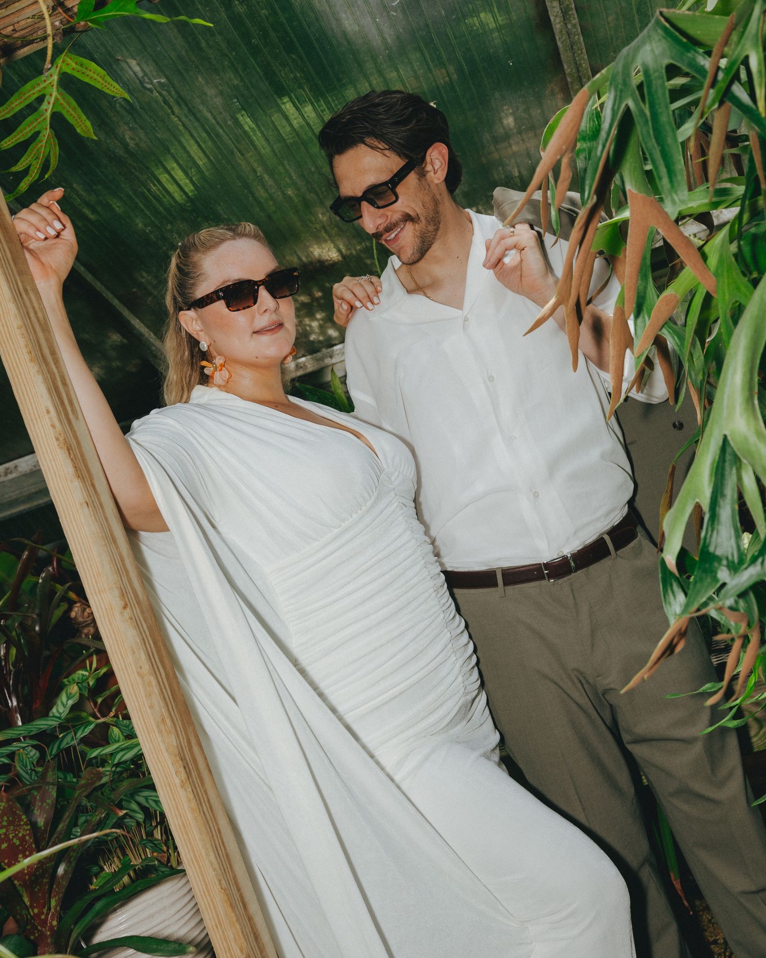 A couple in white wedding attire and sunglasses pose among tropical plants in a greenhouse setting.