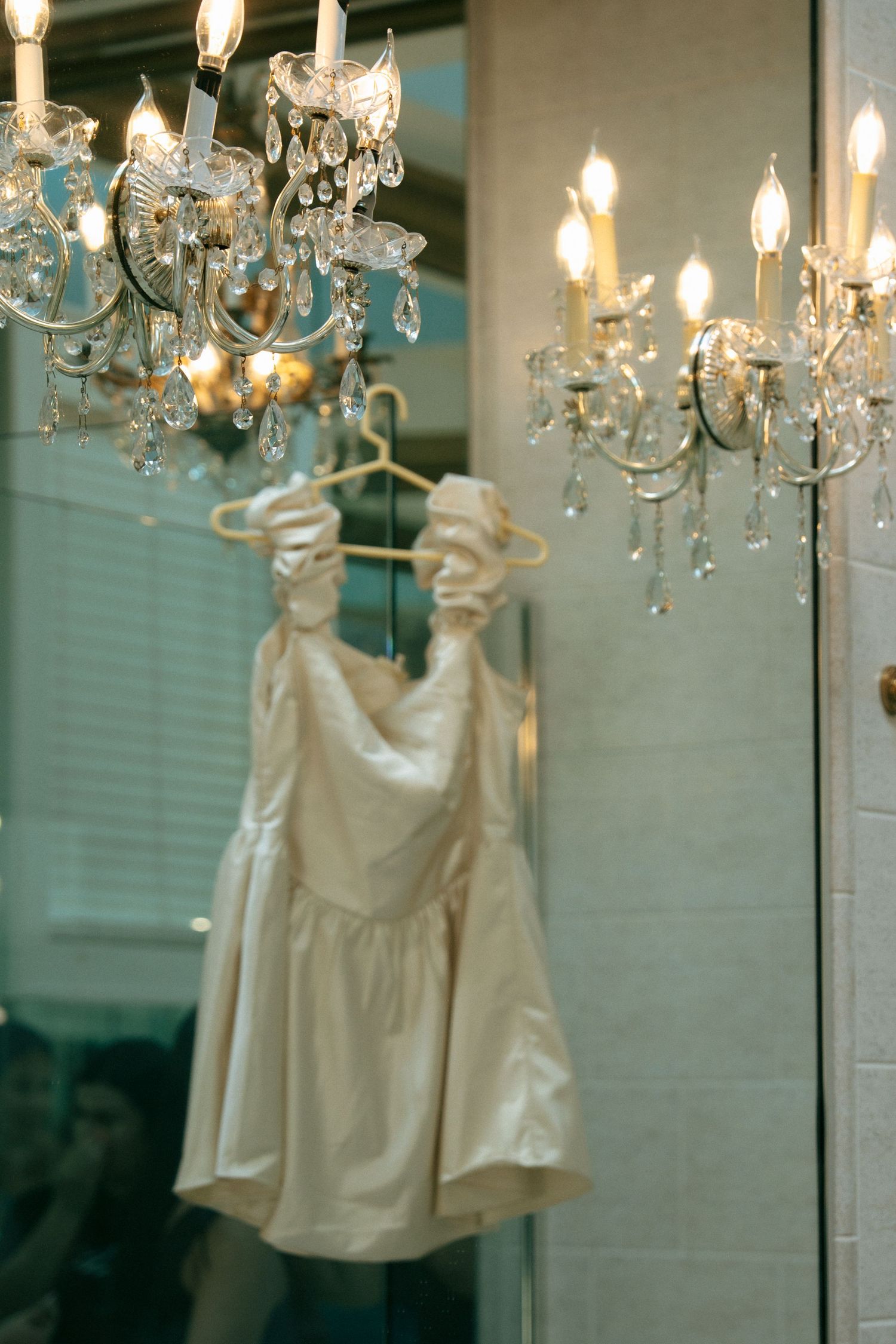 Elegant white satin flower girl dress hanging from a crystal chandelier in a softly lit room.