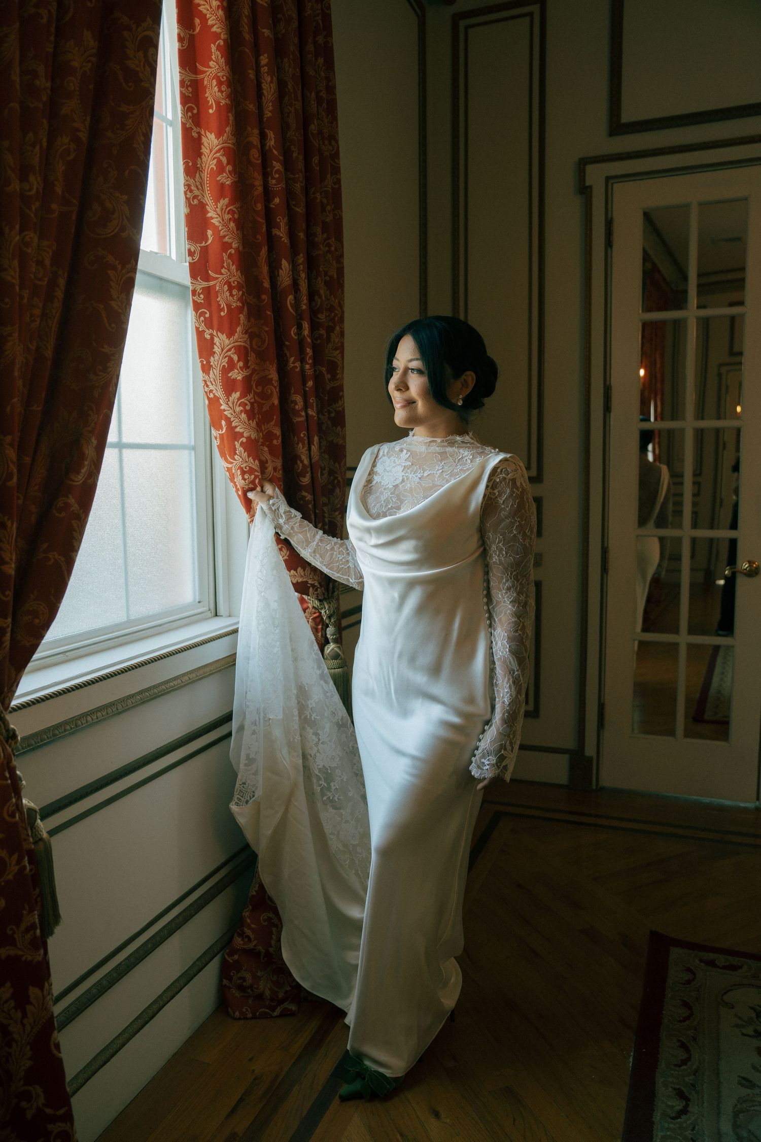 Bride in elegant white lace long-sleeve gown standing by a window with red floral curtains in a classic interior.