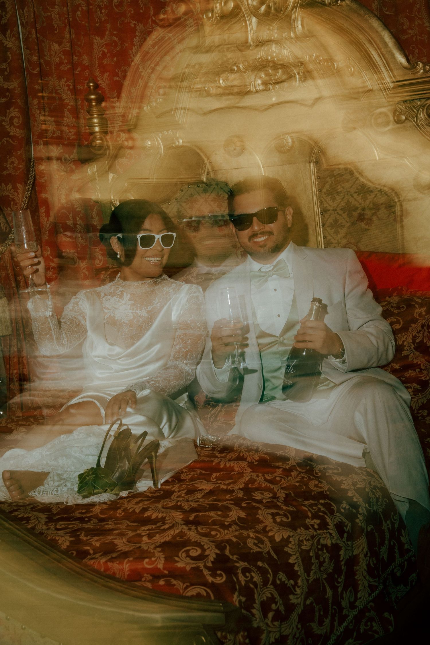 Bride and groom in white outfits and sunglasses celebrate with champagne on ornate red bed in dreamy double exposure photo.