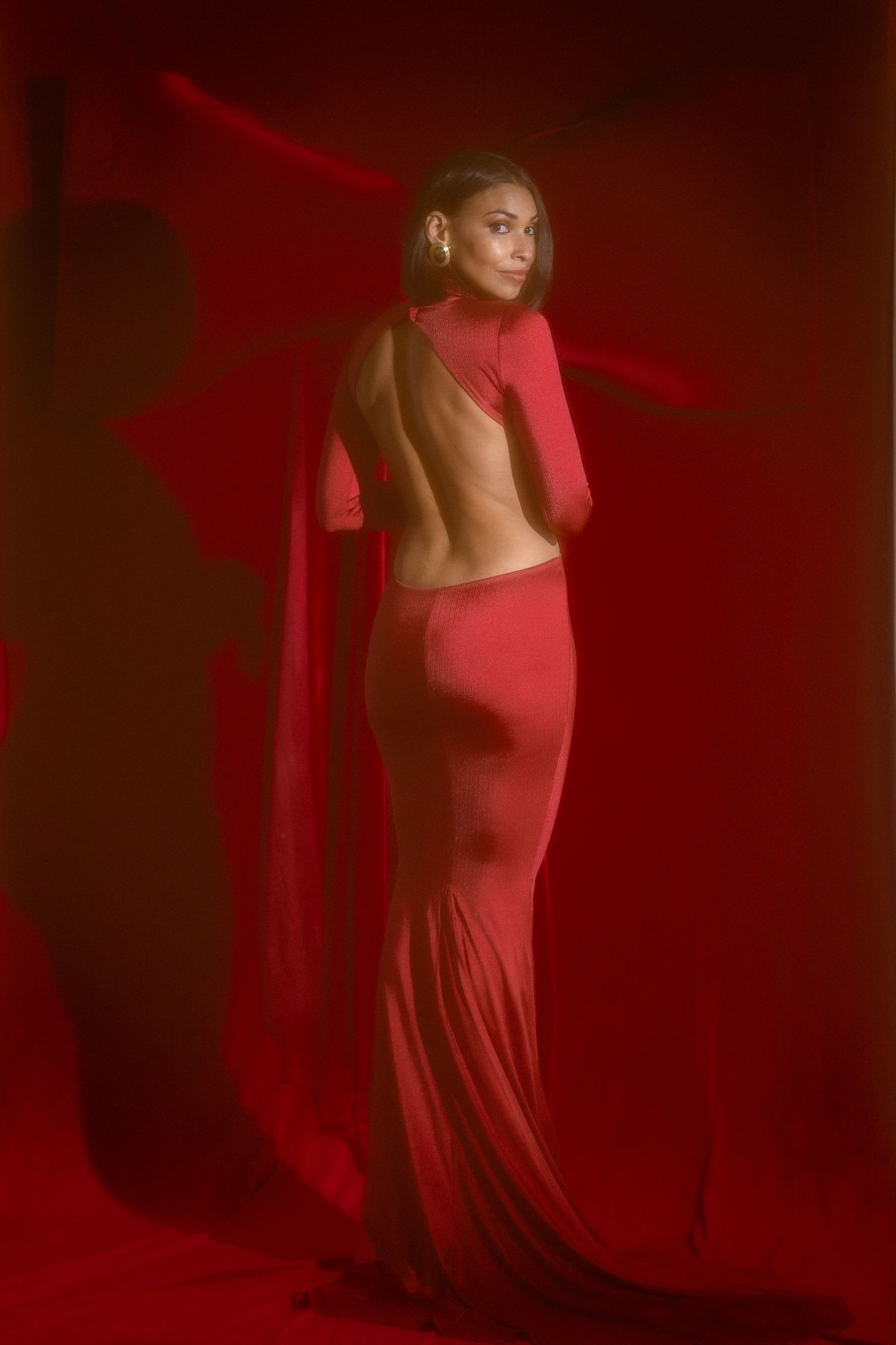 Woman in stunning backless red gown with cape train posing against rich red backdrop.