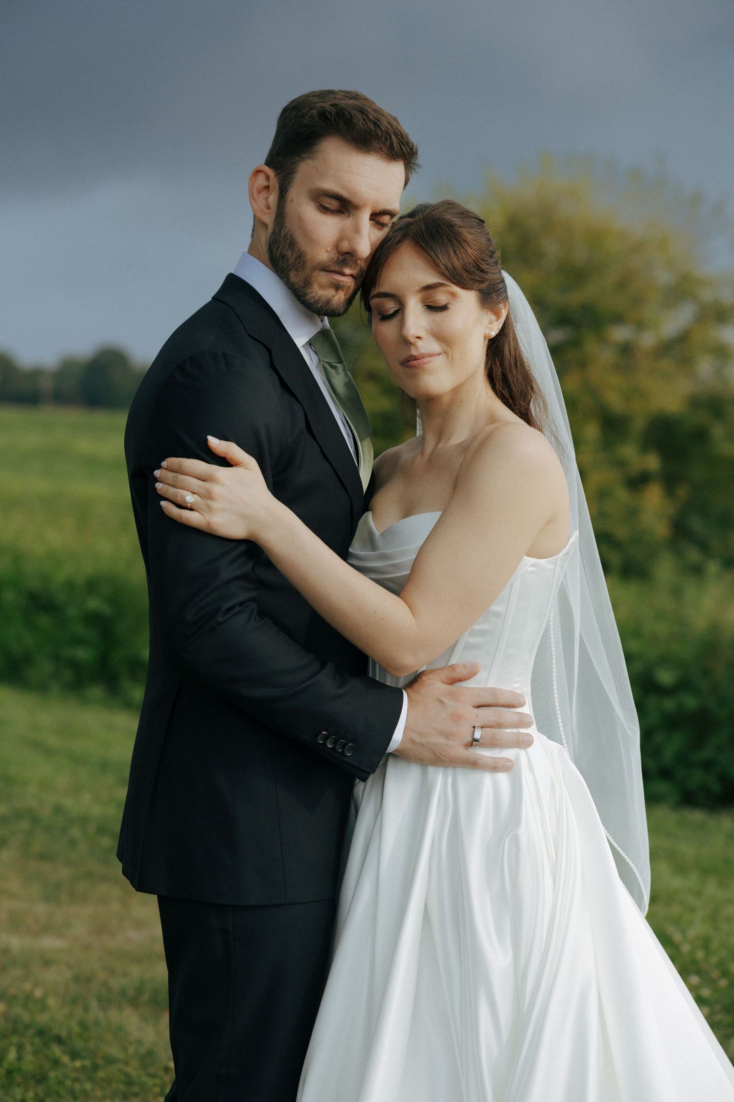 Bride and groom embracing tenderly in a green field under dramatic stormy skies on their wedding day.