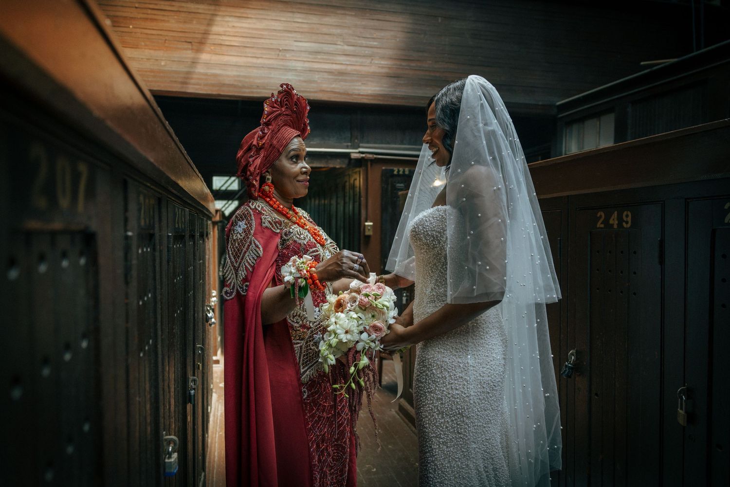 Bride in white lace gown and veil holds bouquet beside woman in red traditional attire in elegant hallway.