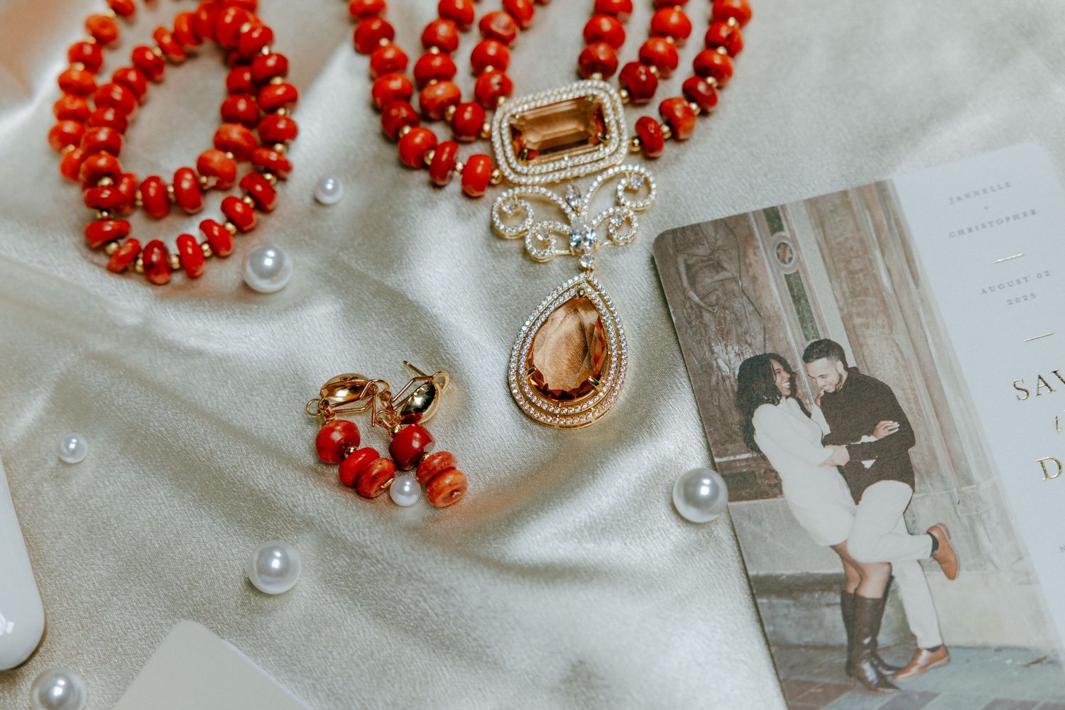 Red coral beaded necklace with gold pendant, earrings, and pearl accents displayed on white satin fabric beside a photo.