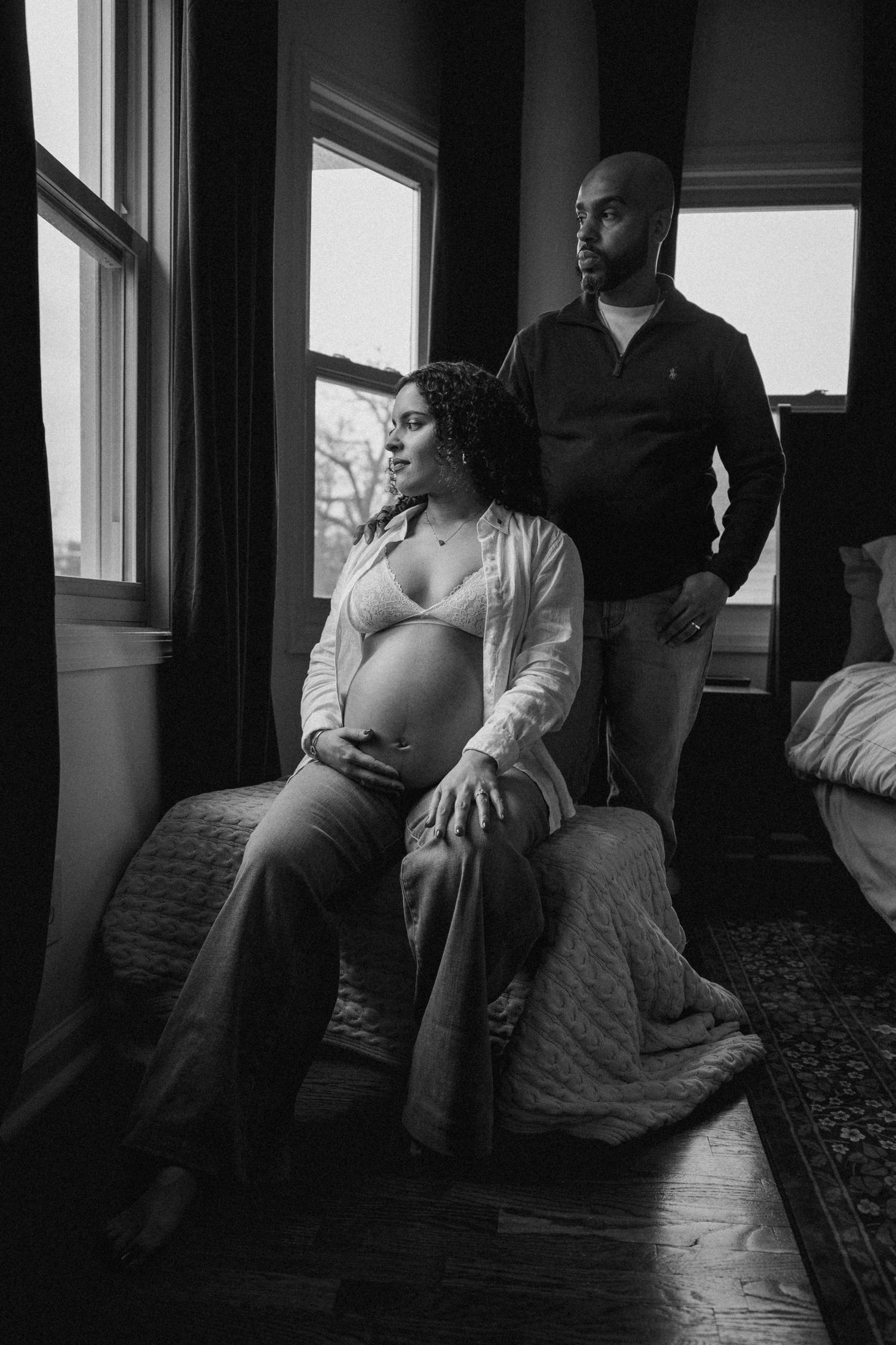Black and white maternity photo of pregnant woman sitting on bed with partner standing behind her near window.