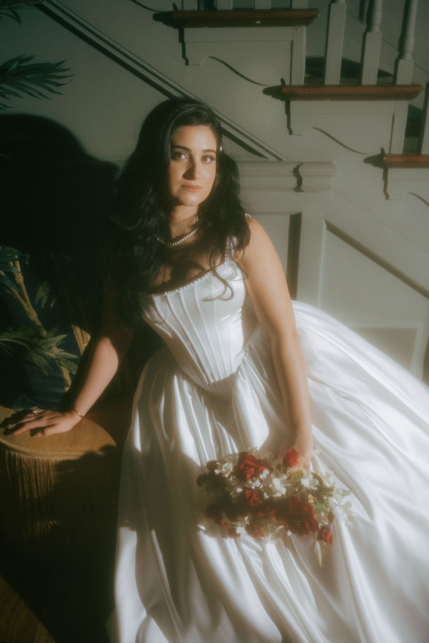 Bride in white corset ballgown holding red and white bouquet, seated by staircase in soft natural light.