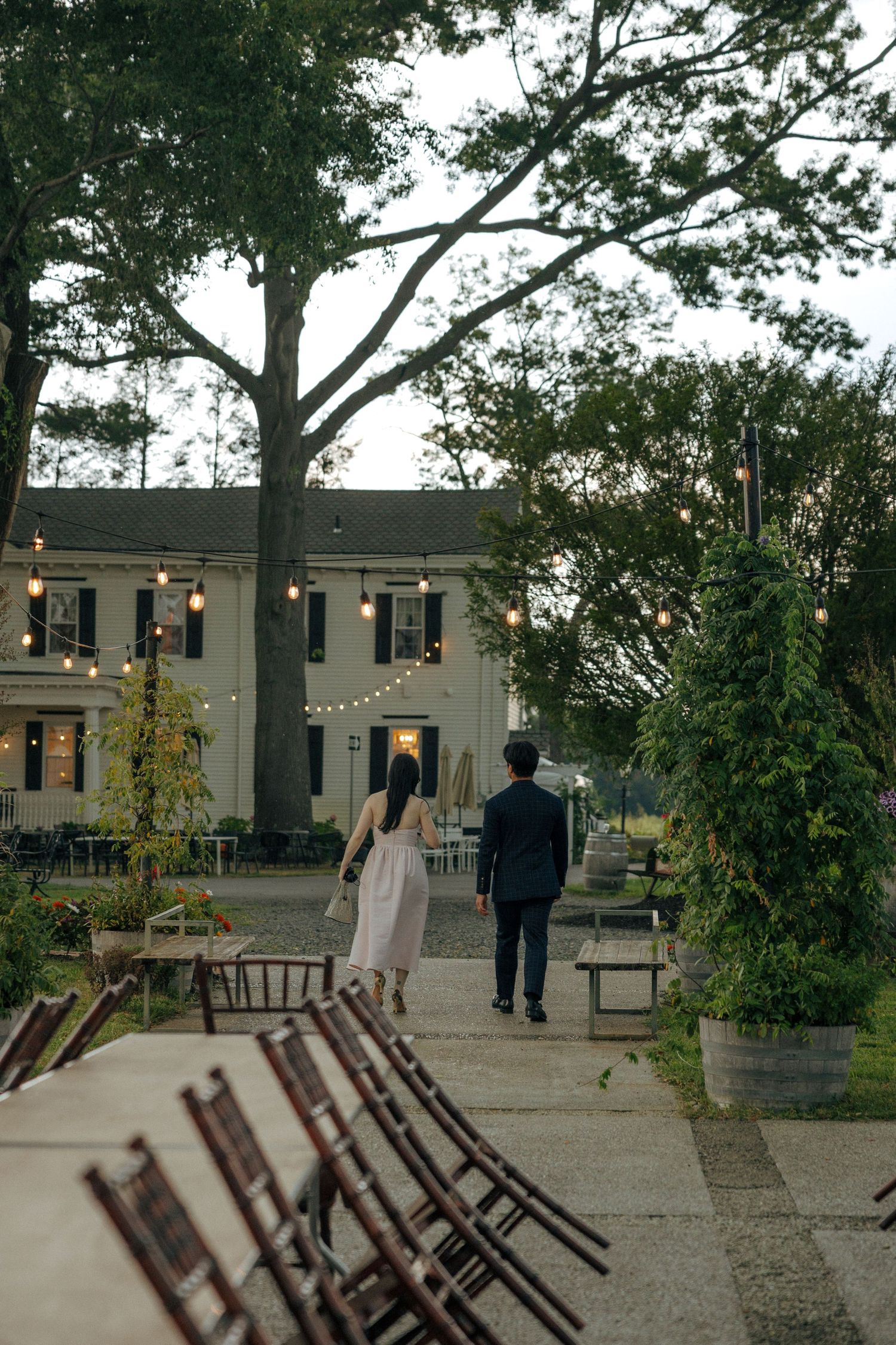 Couple walking toward elegant venue with string lights and lush trees at a romantic outdoor wedding.