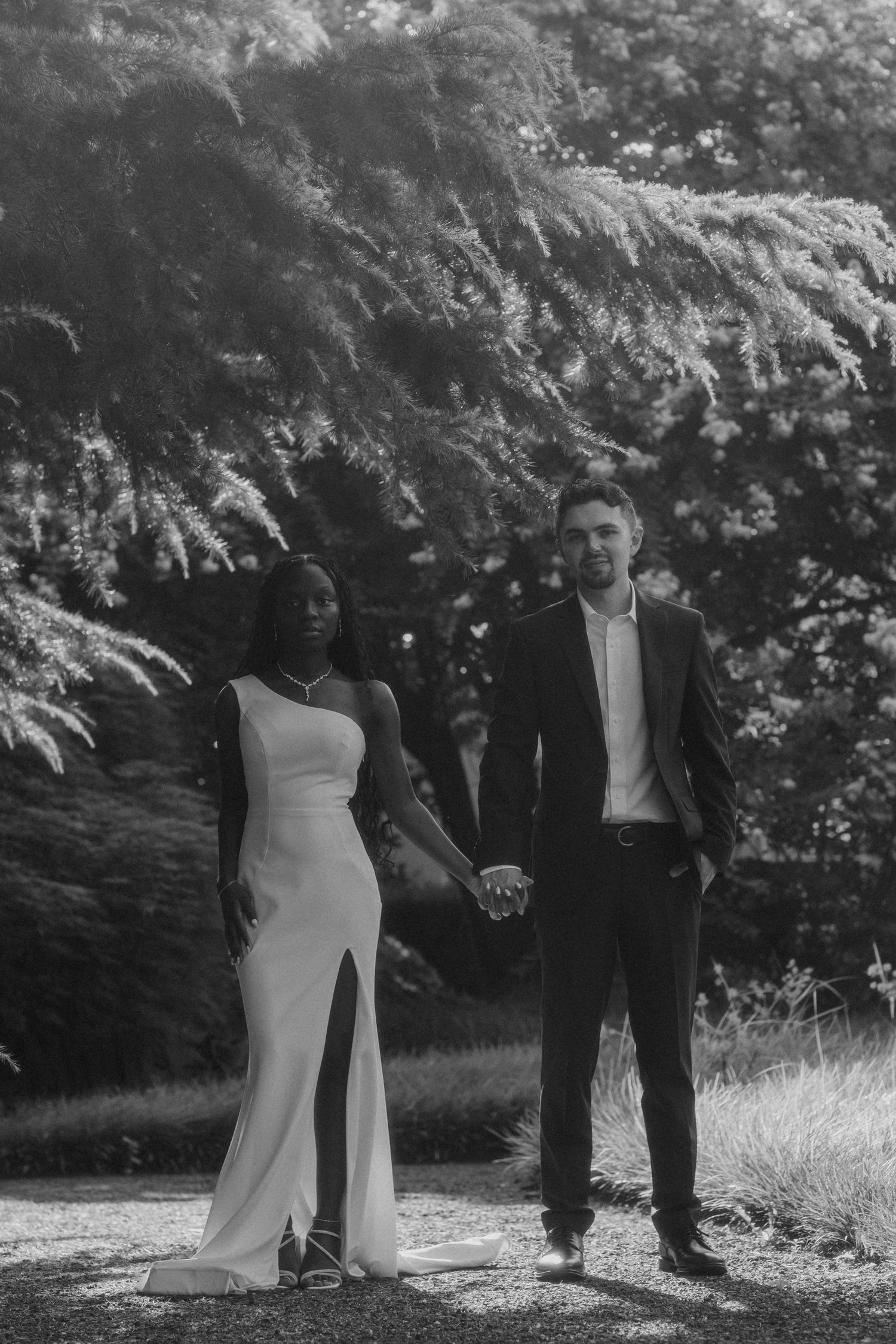 Black and white photo of an interracial couple holding hands in a garden, woman in white gown, man in dark suit.