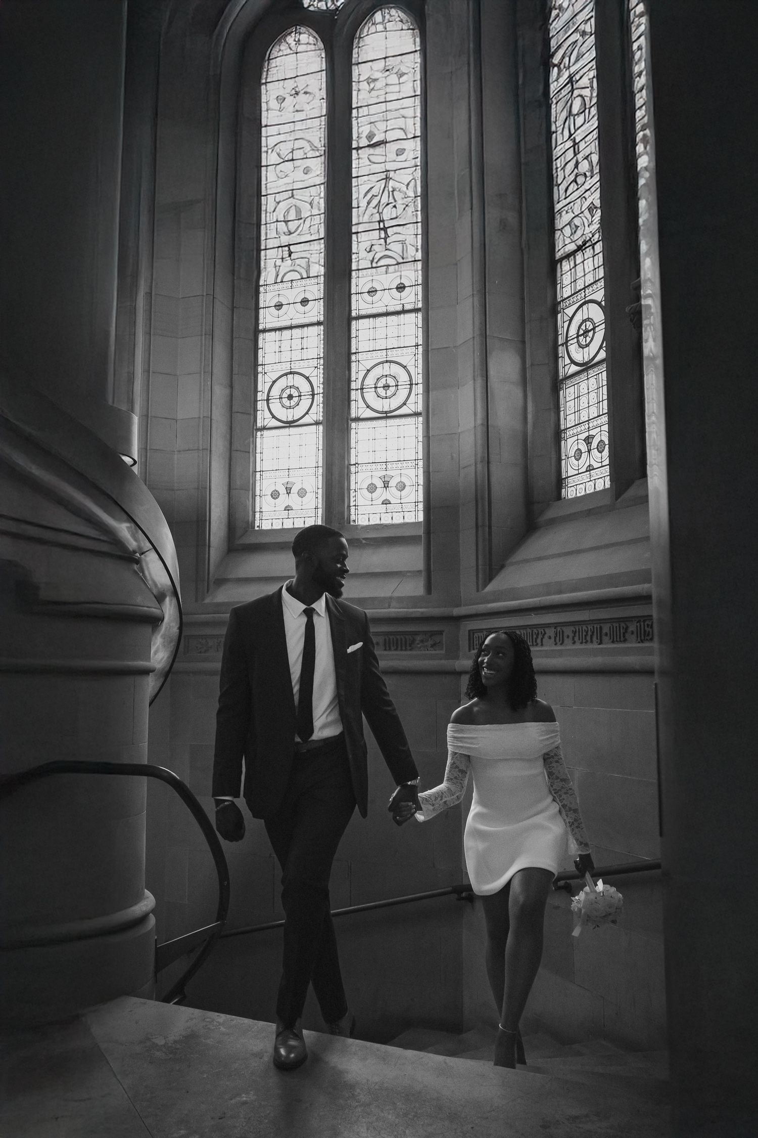 Black and white photo of a couple holding hands walking through a grand hall with stained glass windows.