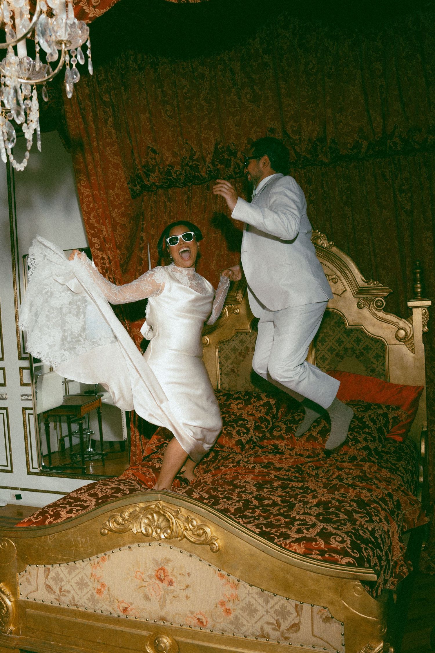 Bride and groom joyfully jumping on an ornate golden bed in a lavish room with chandelier and rich red drapes.