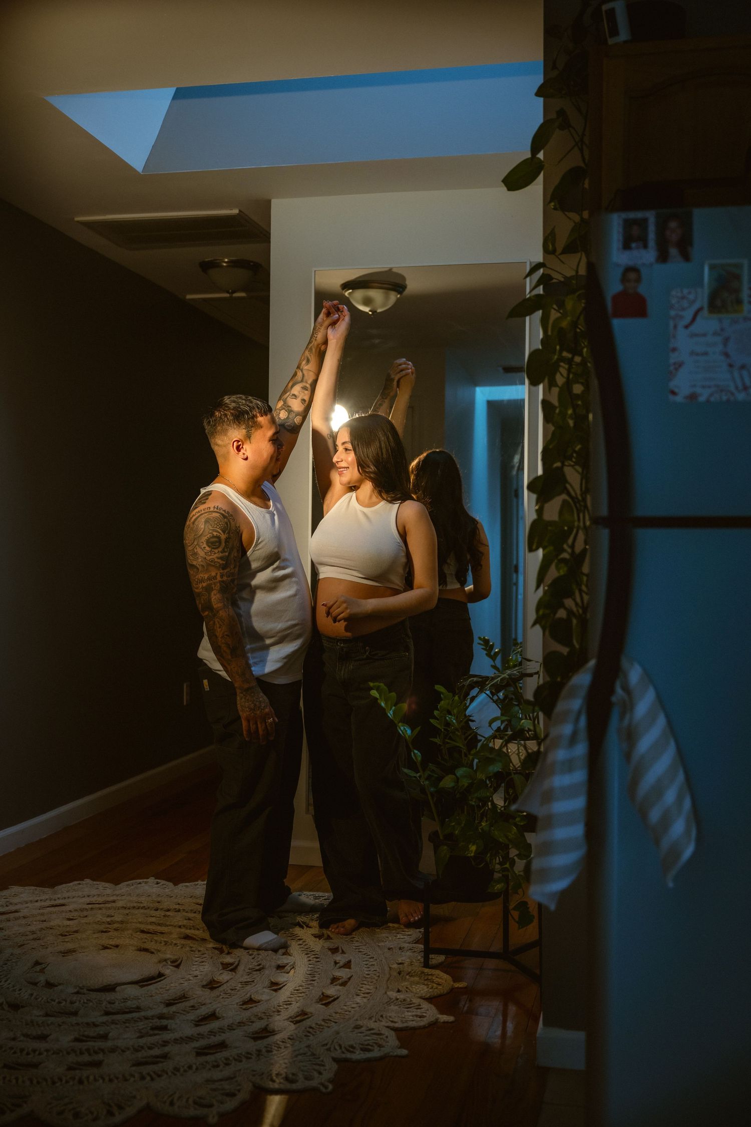 Two people dancing together in a warmly lit home hallway with plants and tattoos visible.