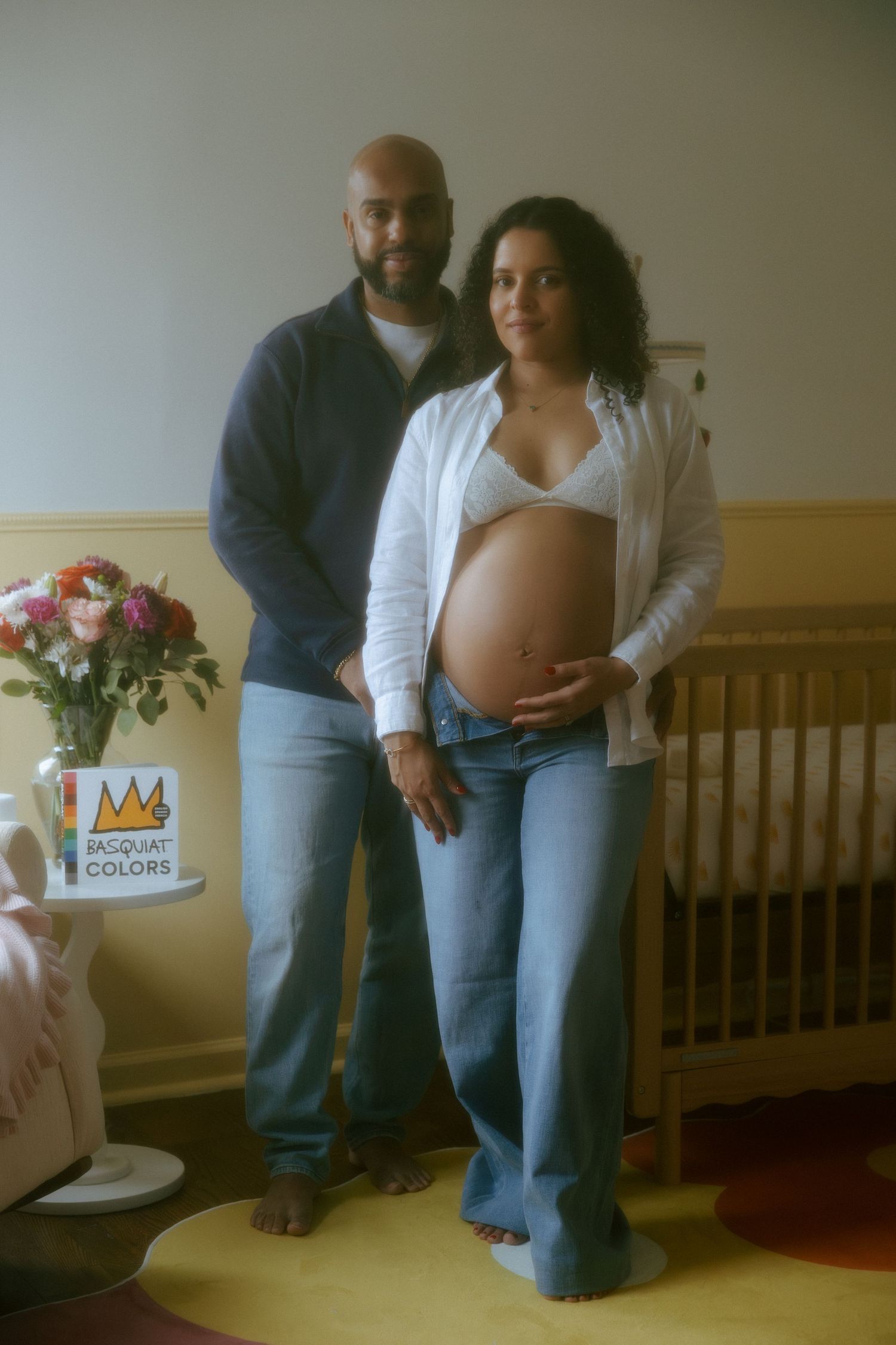 Pregnant woman in white top and jeans poses with partner in nursery room featuring a crib and Basquiat Colors book.