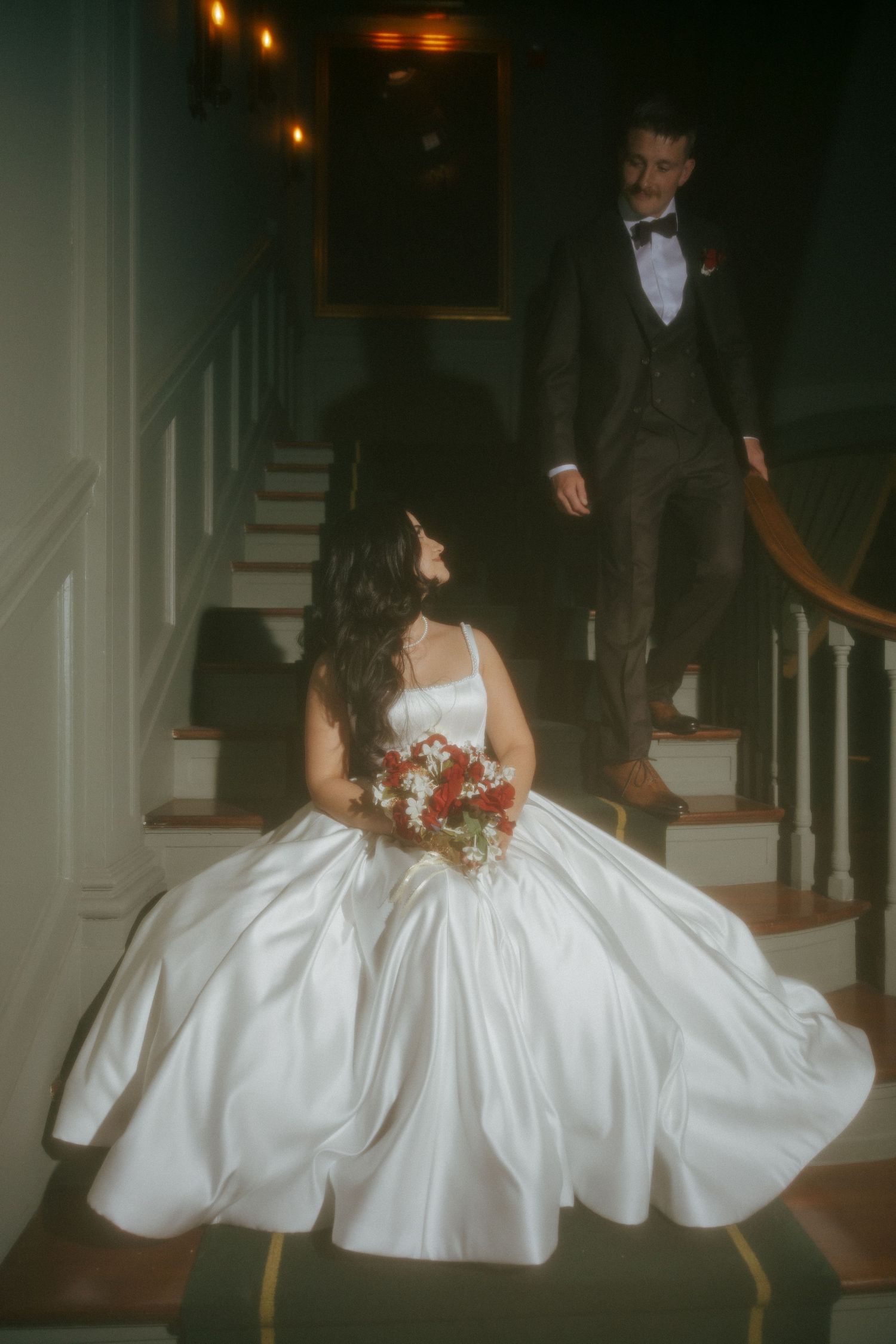 Bride in white ballgown sits on staircase holding red bouquet while groom in dark suit descends toward her.
